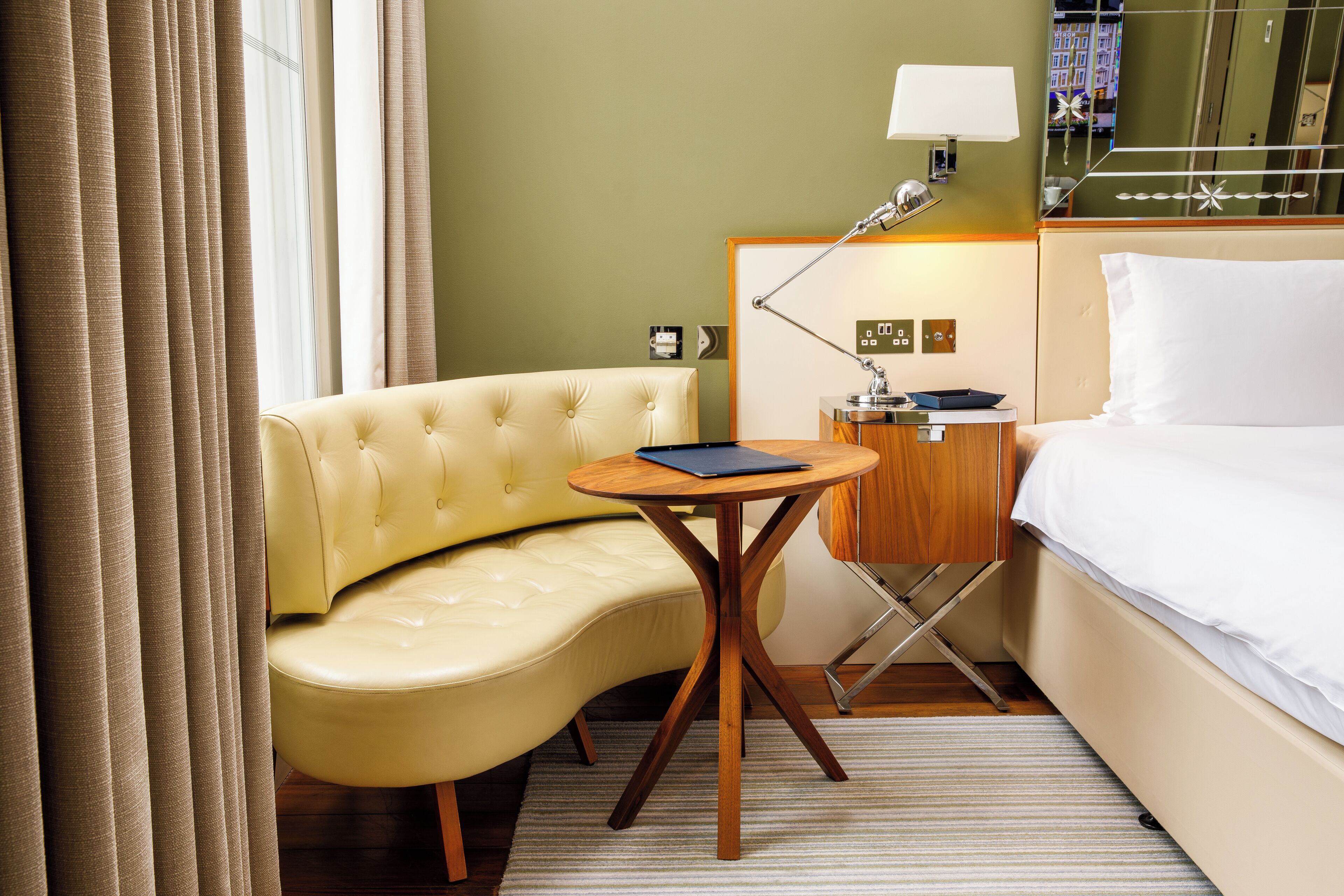 Habitación de hotel con sofá amarillo curvo, una pequeña mesa de madera, lámpara de noche y cama con ropa blanca.