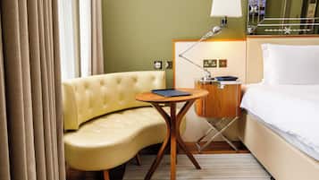 Habitación de hotel con sofá amarillo curvo, una pequeña mesa de madera, lámpara de noche y cama con ropa blanca.