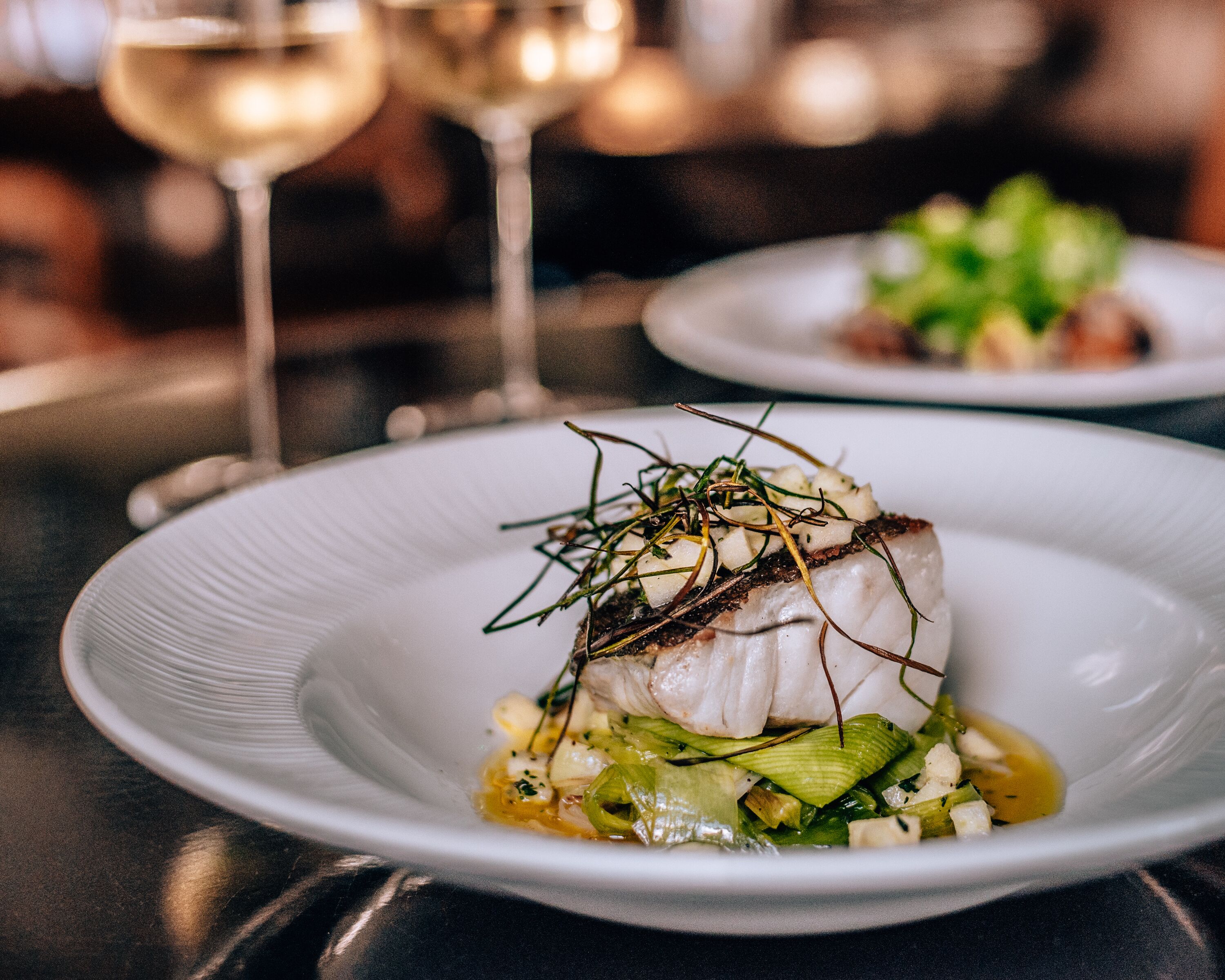 Ein weißer Teller mit einem Fischgericht, garniert mit Kräutern und Gemüse, auf einem Tisch.