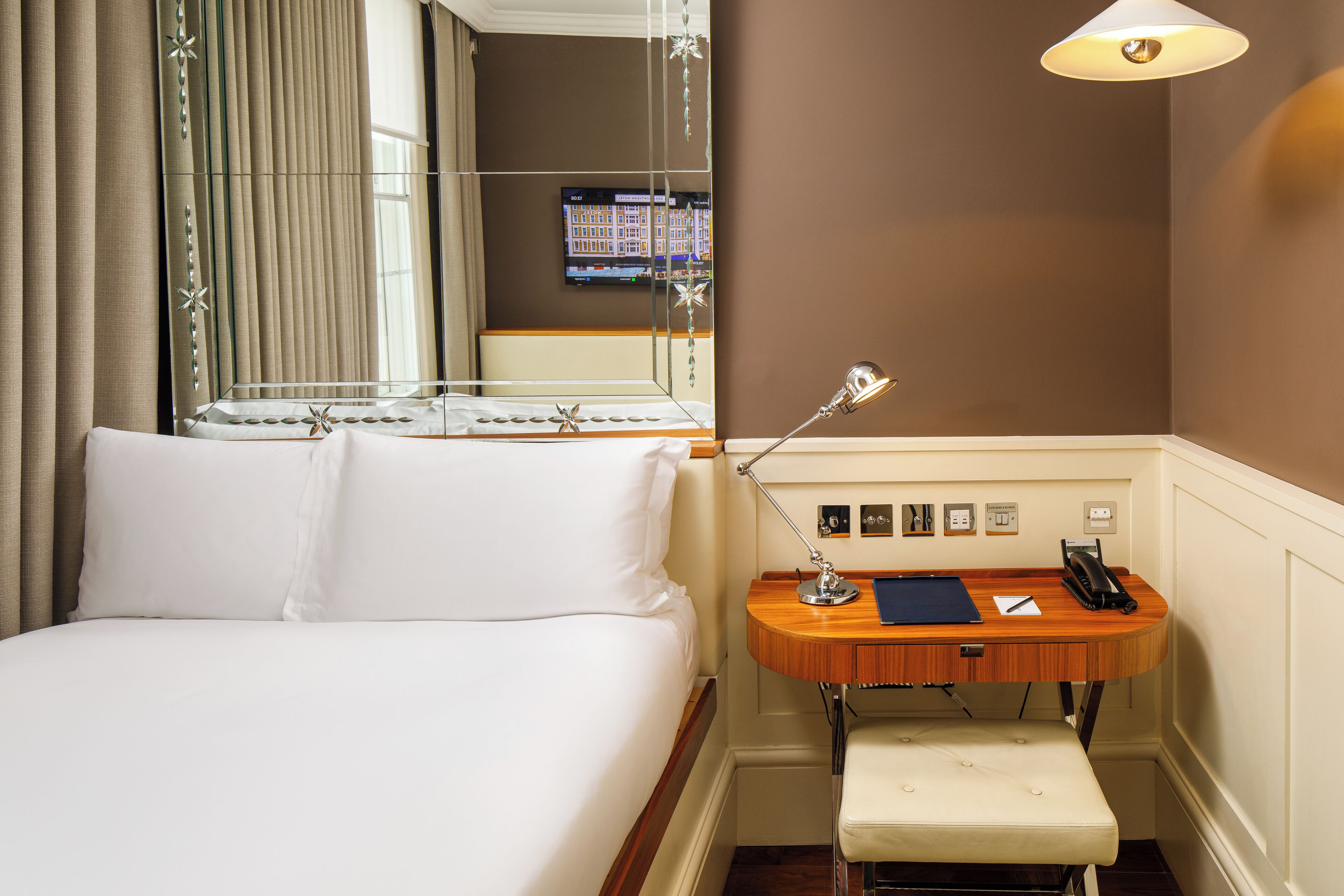A hotel room with a large bed, a wooden desk with a lamp, a telephone, and a small bench.