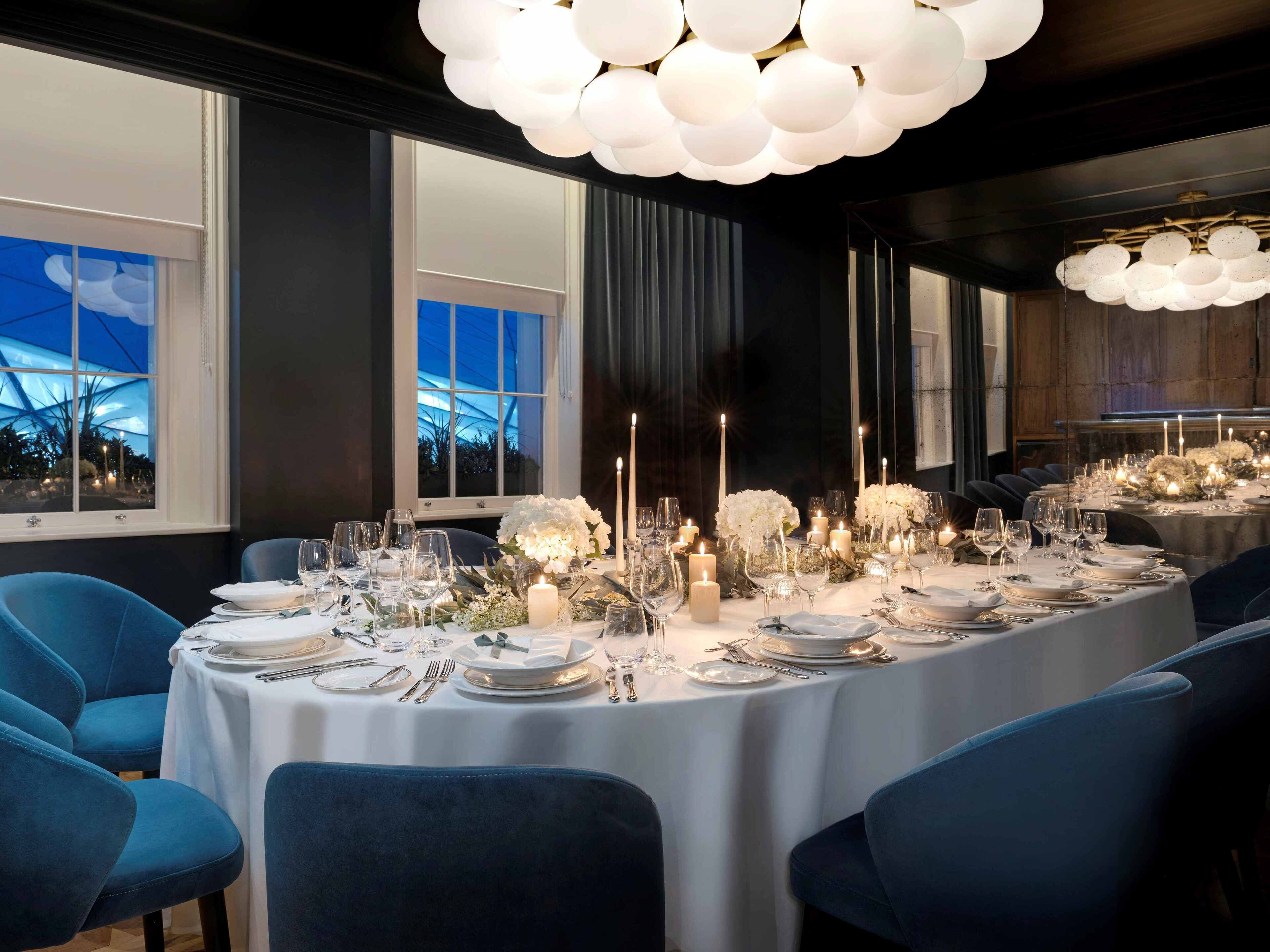 Ein formeller Speisesaal mit einem runden Tisch, gedeckt für eine Mahlzeit, blauen Stühlen und einer modernen Kronleuchter.