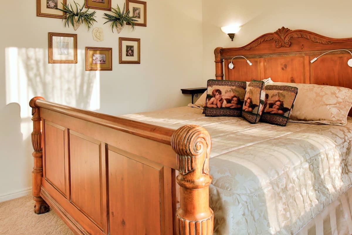 A wooden bed with a headboard, a bedside table, and a wall with framed pictures and a plant.