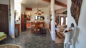 A spacious living area with a dining table, chairs, and a sofa. There is a kitchen area with cabinets and a chandelier. The room has a tiled floor and a large window.