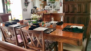 A dining area with a wooden table set for four, surrounded by wooden chairs with floral cushions. A potted plant with white flowers is on the table.
