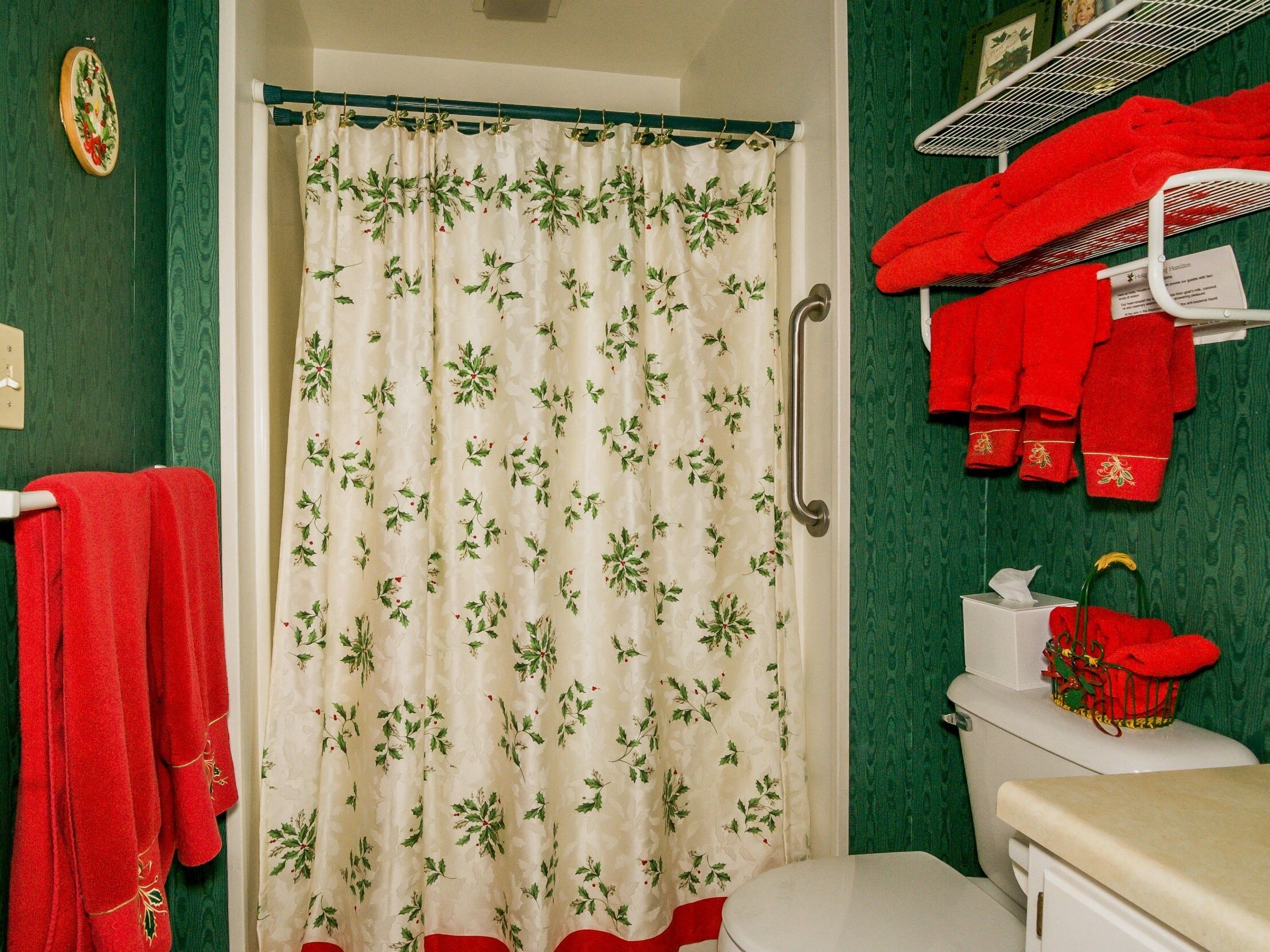 A bathroom with a shower curtain featuring a holly pattern, a white toilet, and red towels hanging on a rack.