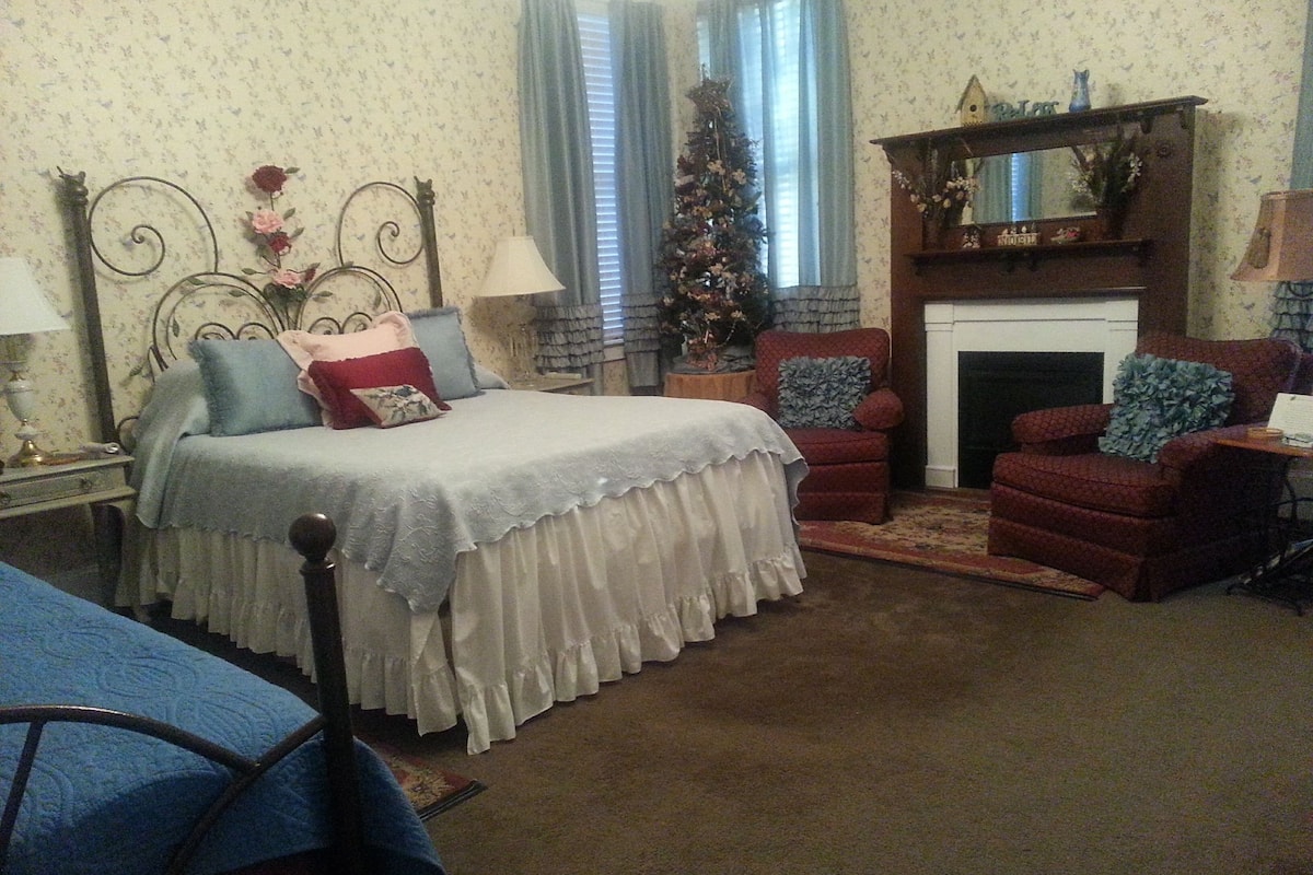 A bedroom with a bed, two chairs, a fireplace, and a Christmas tree.