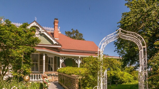 Een huis in Victoriaanse stijl met een veranda, een houten pad en een decoratieve metalen boog.