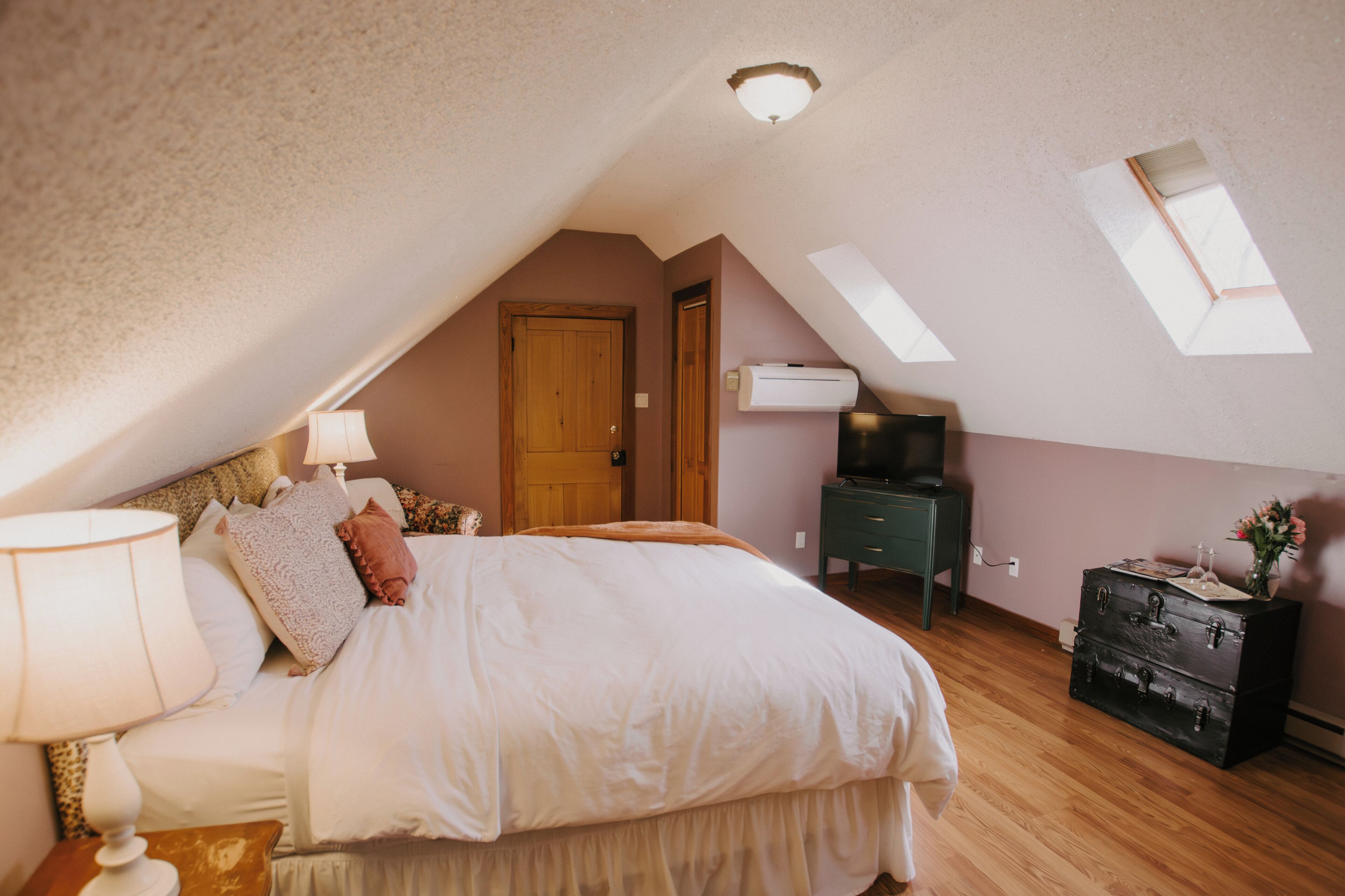 A bedroom with a sloped ceiling, a bed with pillows, a nightstand with a lamp, a dresser, and a television.