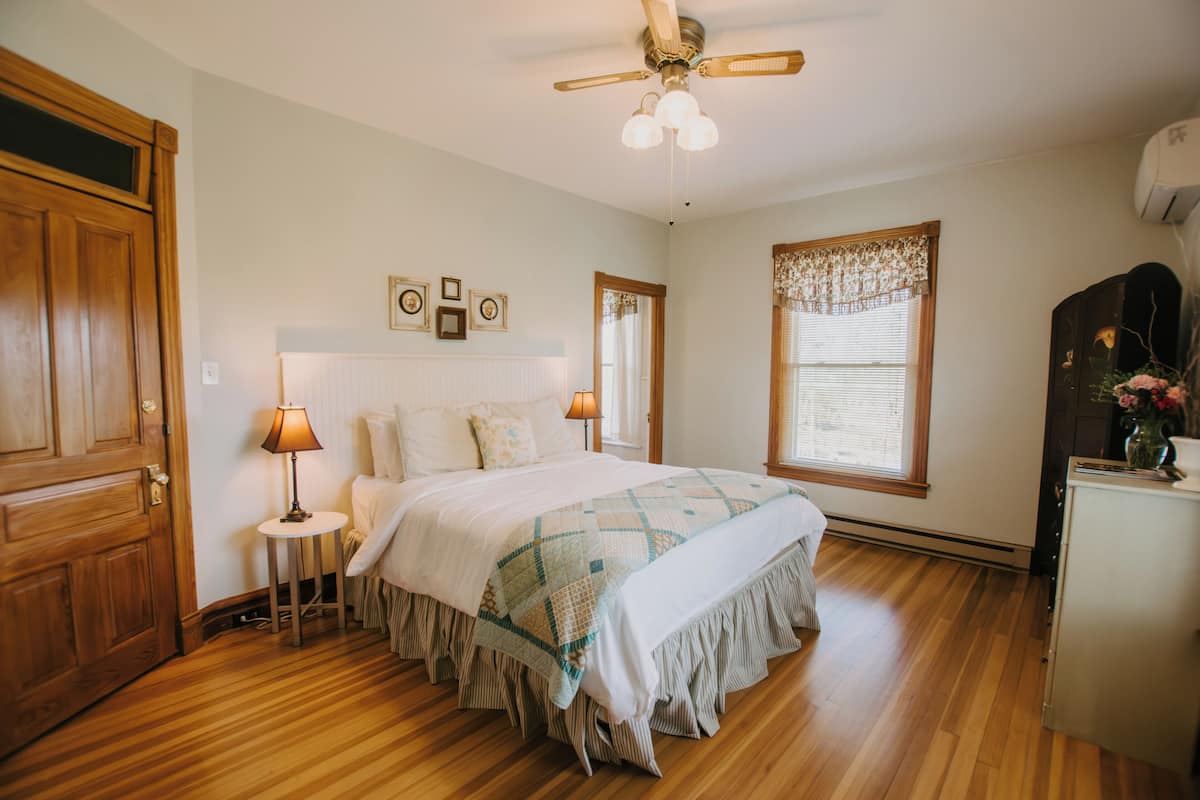 A bedroom with a wooden floor, a bed with a quilt, a ceiling fan, and a window with blinds.