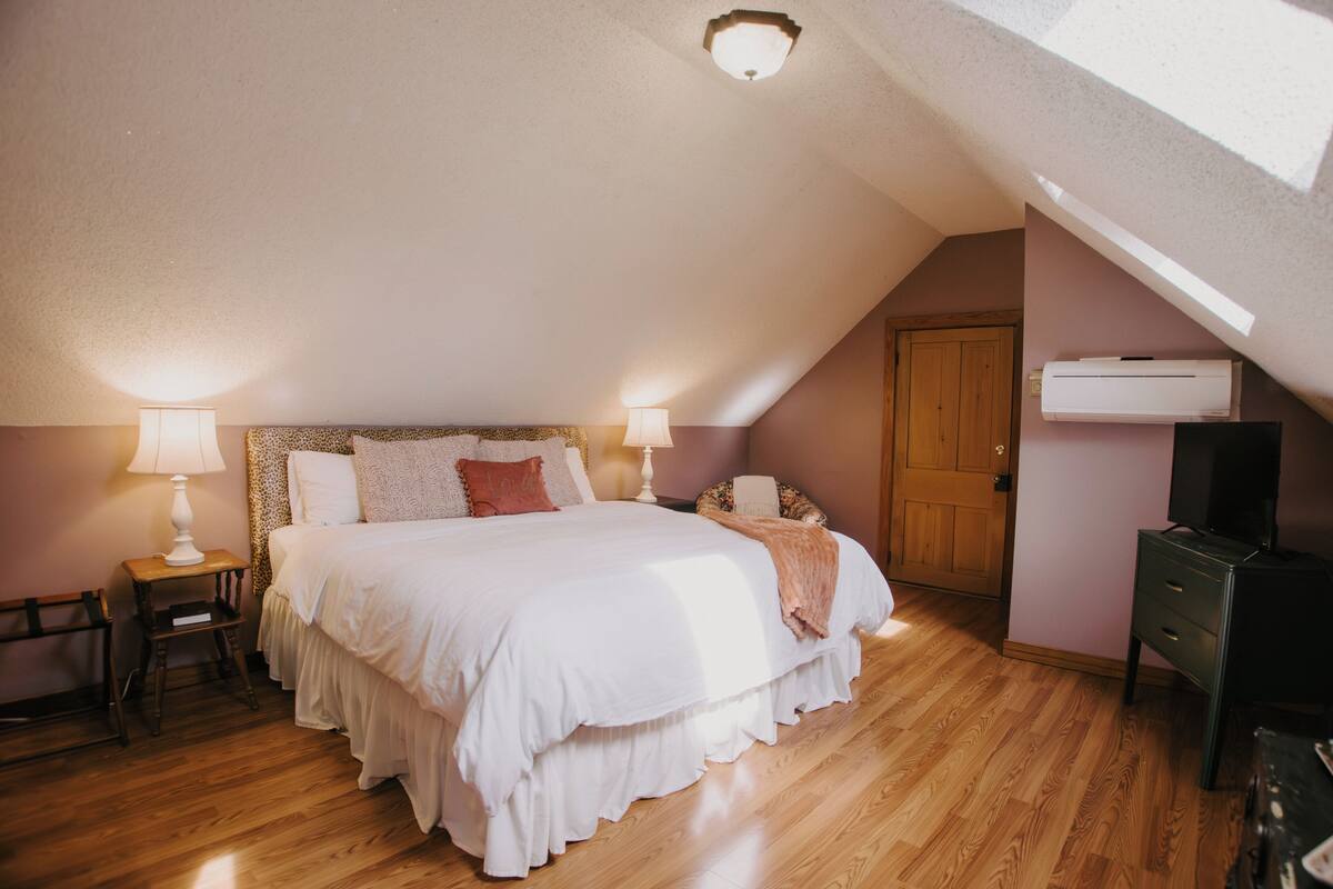 A bedroom with a sloped ceiling, a bed with white bedding, two bedside lamps, a wooden floor, a wardrobe, and an air conditioning unit.