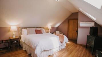 A bedroom with a sloped ceiling, a bed with white bedding, two bedside lamps, a wooden floor, a wardrobe, and an air conditioning unit.