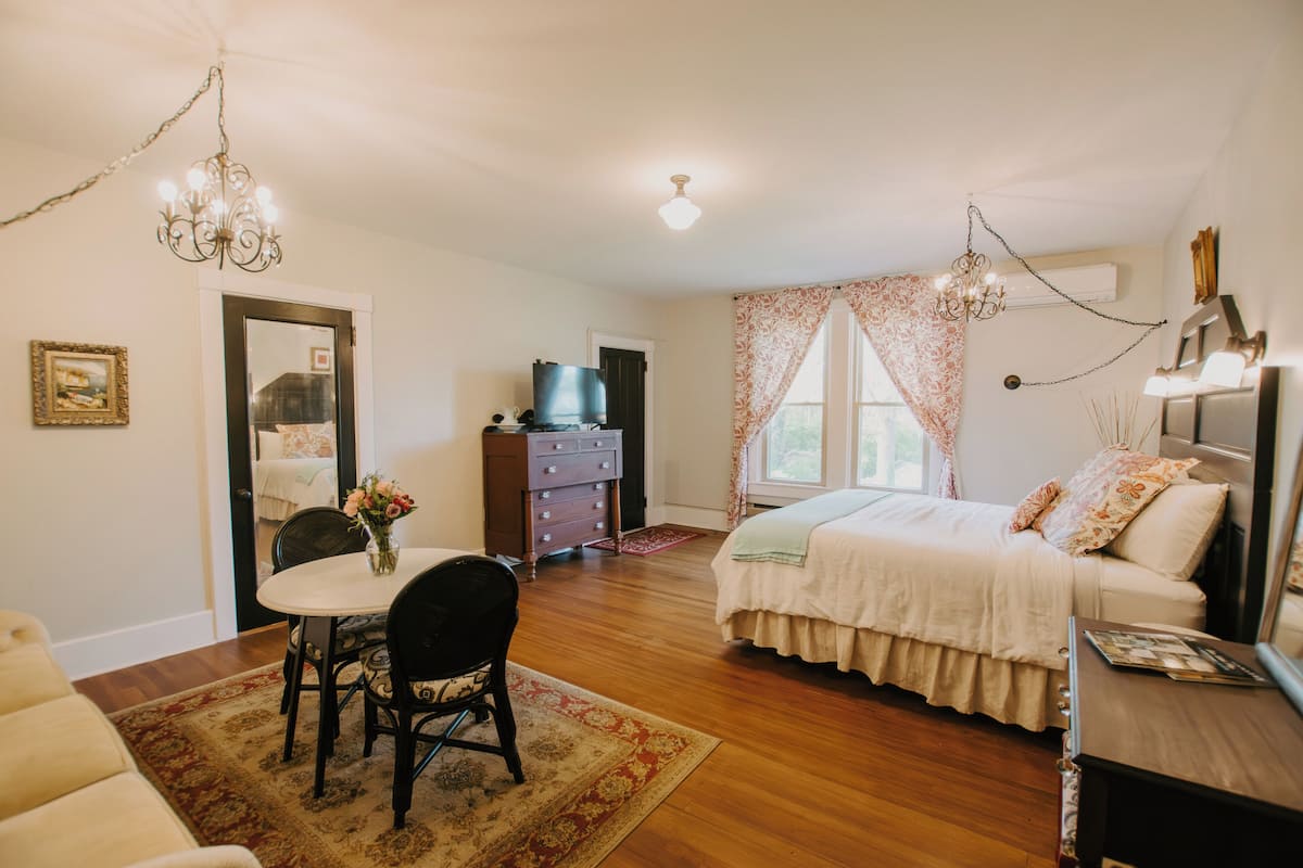 A bedroom with a bed, a dresser, a television, a dining table with chairs, and a chandelier.