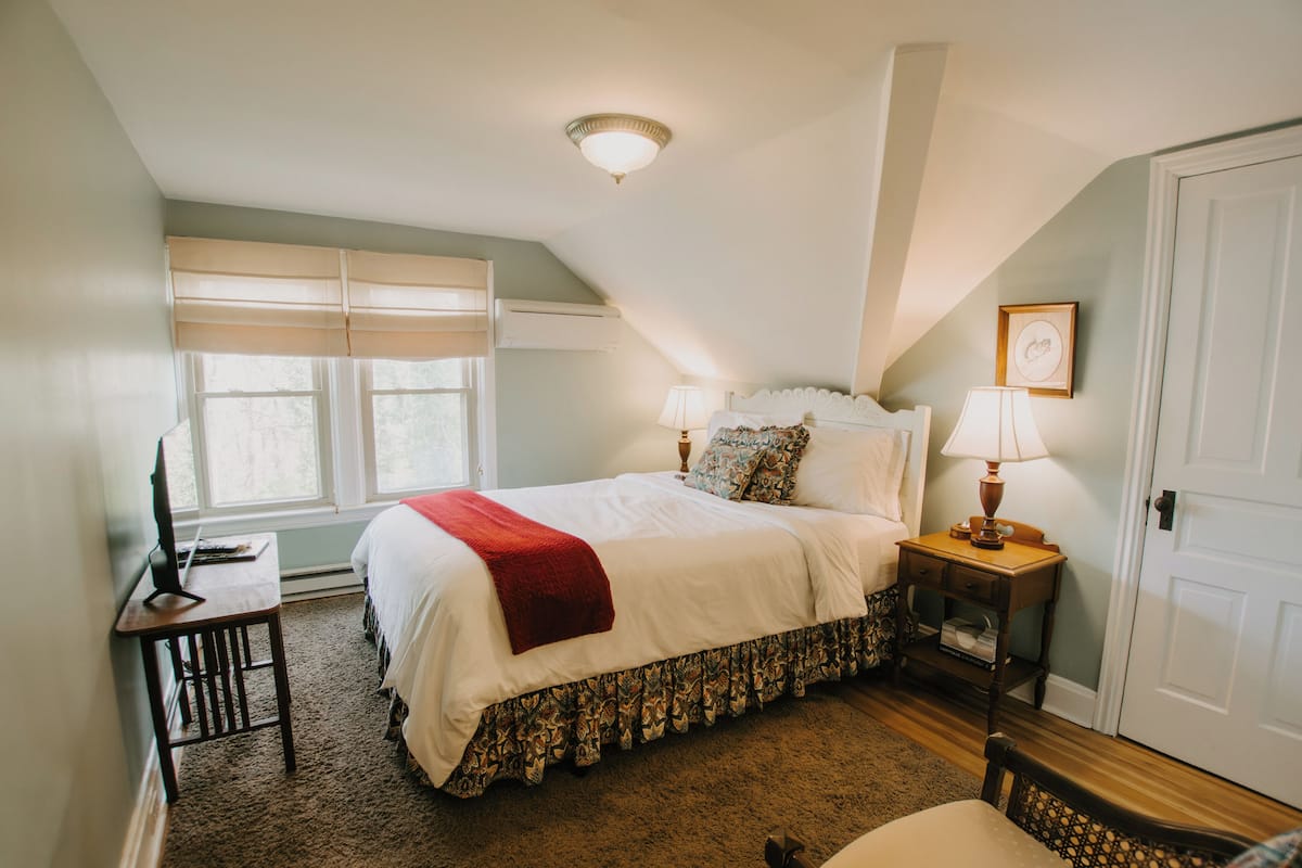 A bedroom with a bed, bedside tables, a desk, a chair, a window with blinds, and a door.
