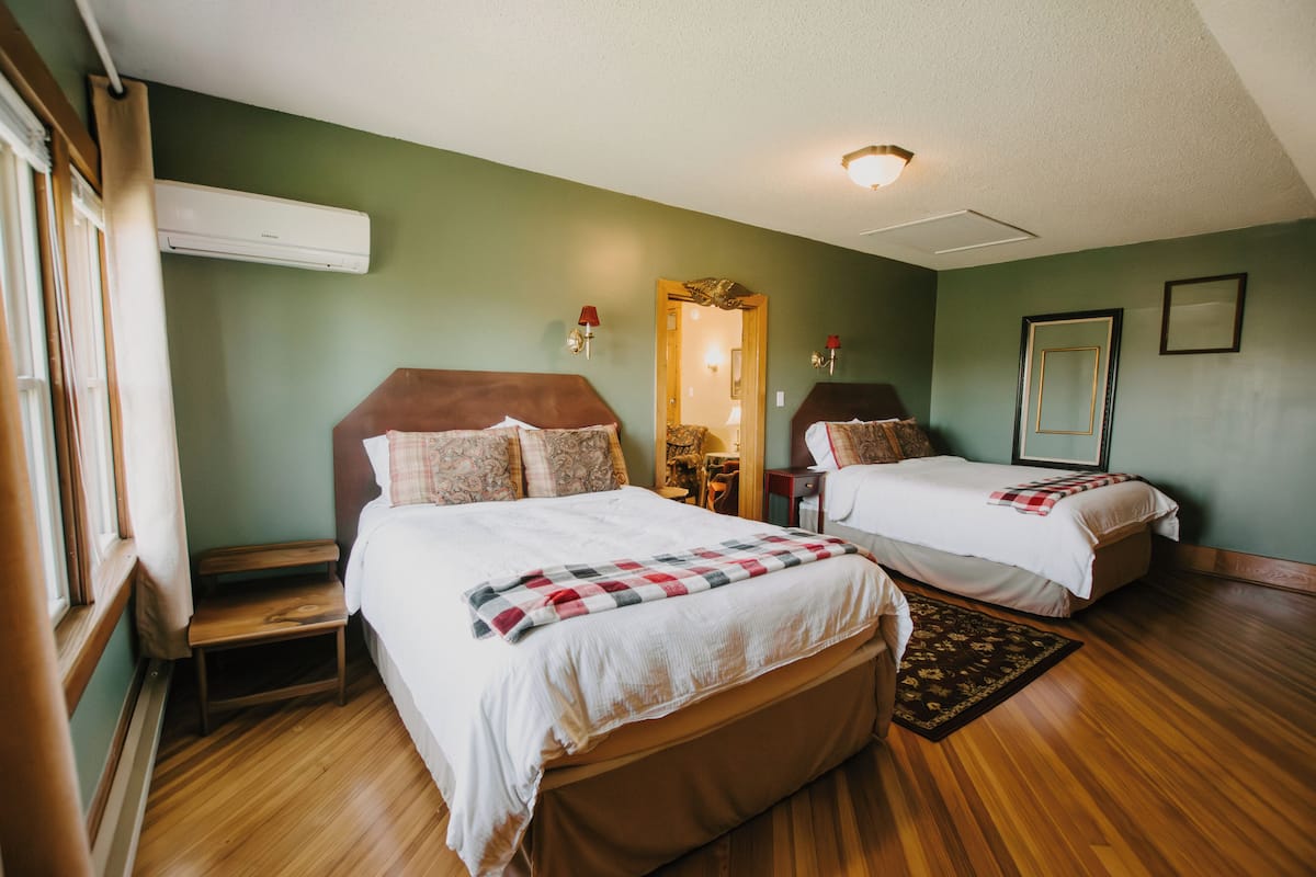 A hotel room with two beds, a wooden floor, a window with blinds, an air conditioner, and a mirror on the wall.