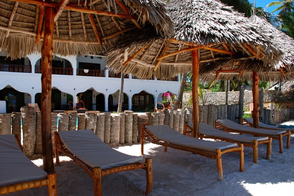 A beachside resort with thatched-roof structures, lounge chairs, and a white building with arched windows.