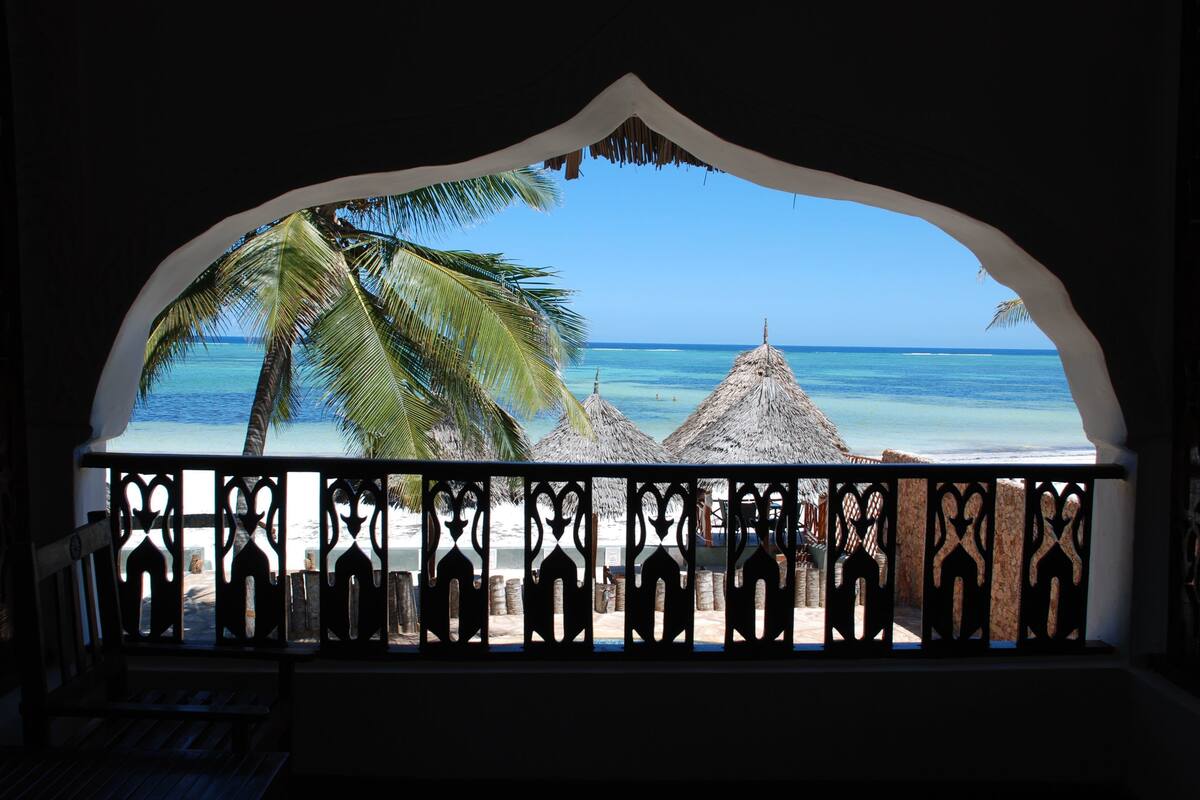 A view from a covered balcony with a thatched roof, overlooking a beach with palm trees and thatched huts.