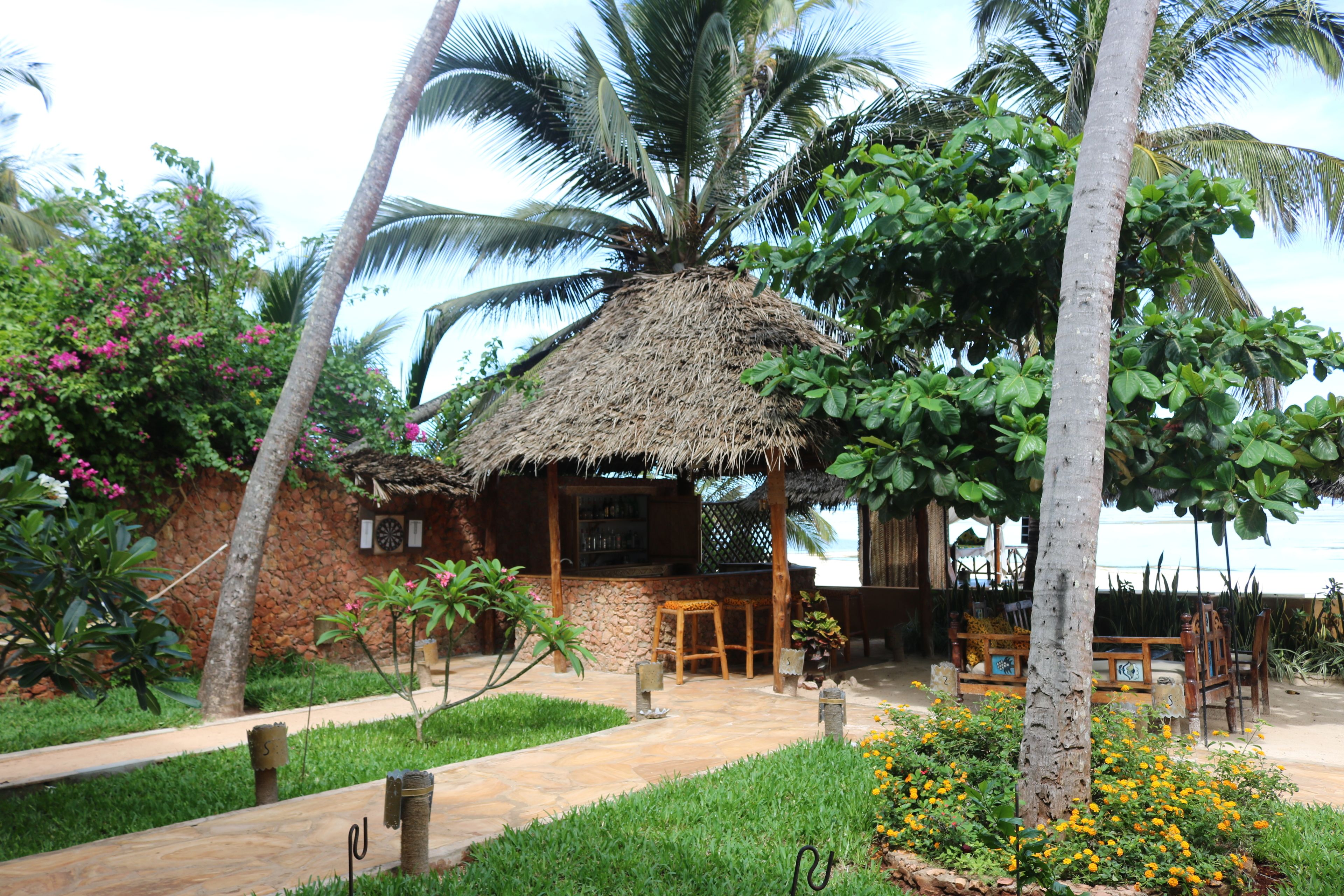 A thatched-roof hut with a stone wall, surrounded by palm trees and a garden with yellow flowers.