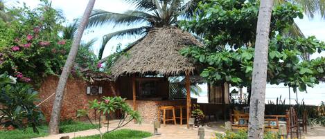 A thatched-roof hut with a stone wall, surrounded by palm trees and a garden with yellow flowers.