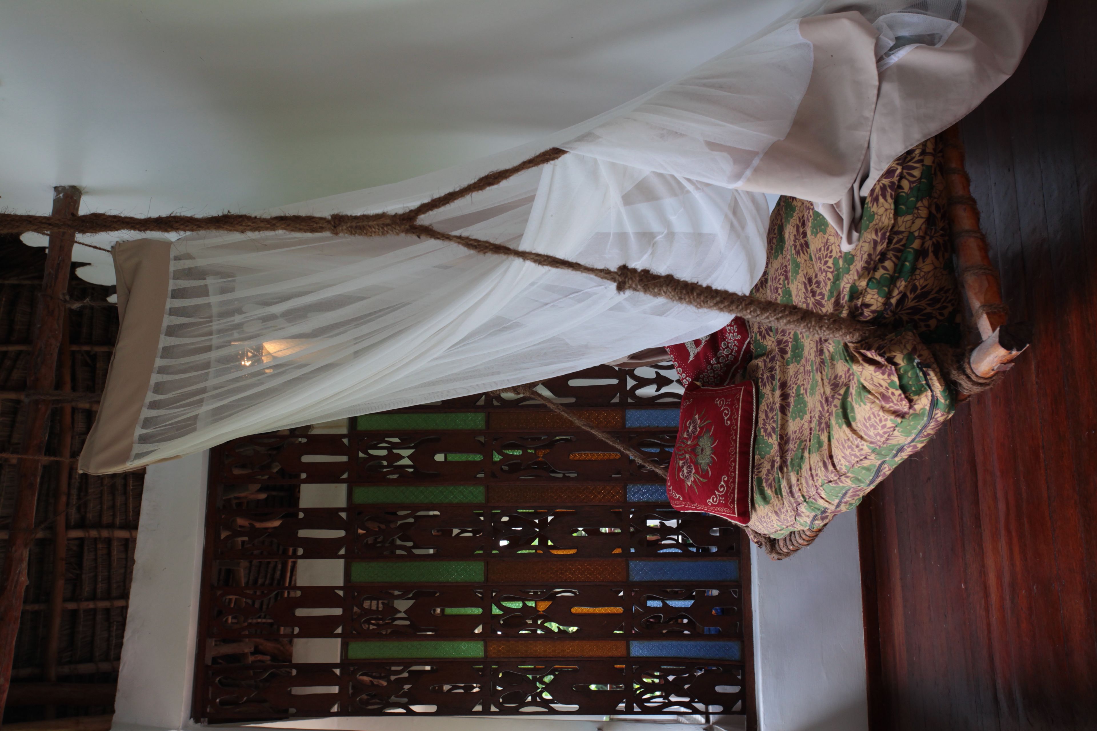 A room with a bed covered by a white canopy, a wooden lattice partition, and a red patterned bedspread.