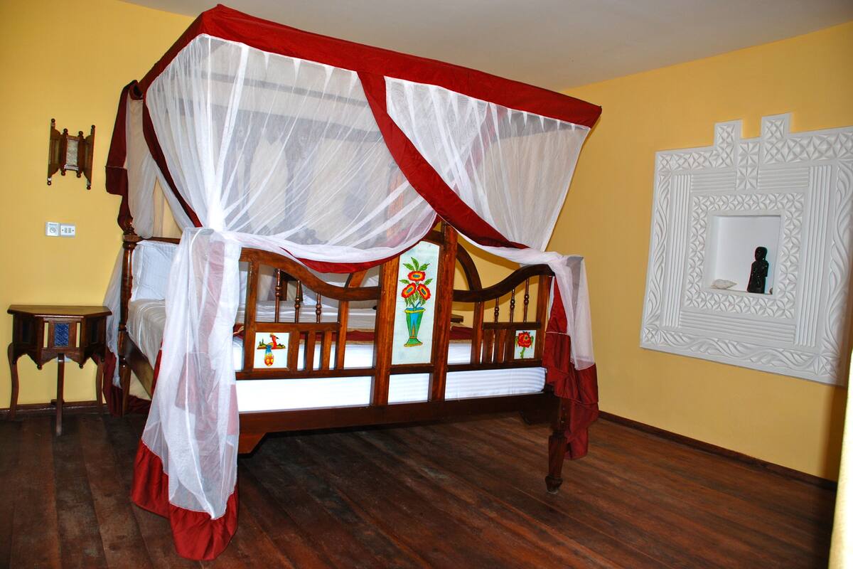 A four-poster bed with a canopy, wooden flooring, a small table, and a framed picture on the wall.