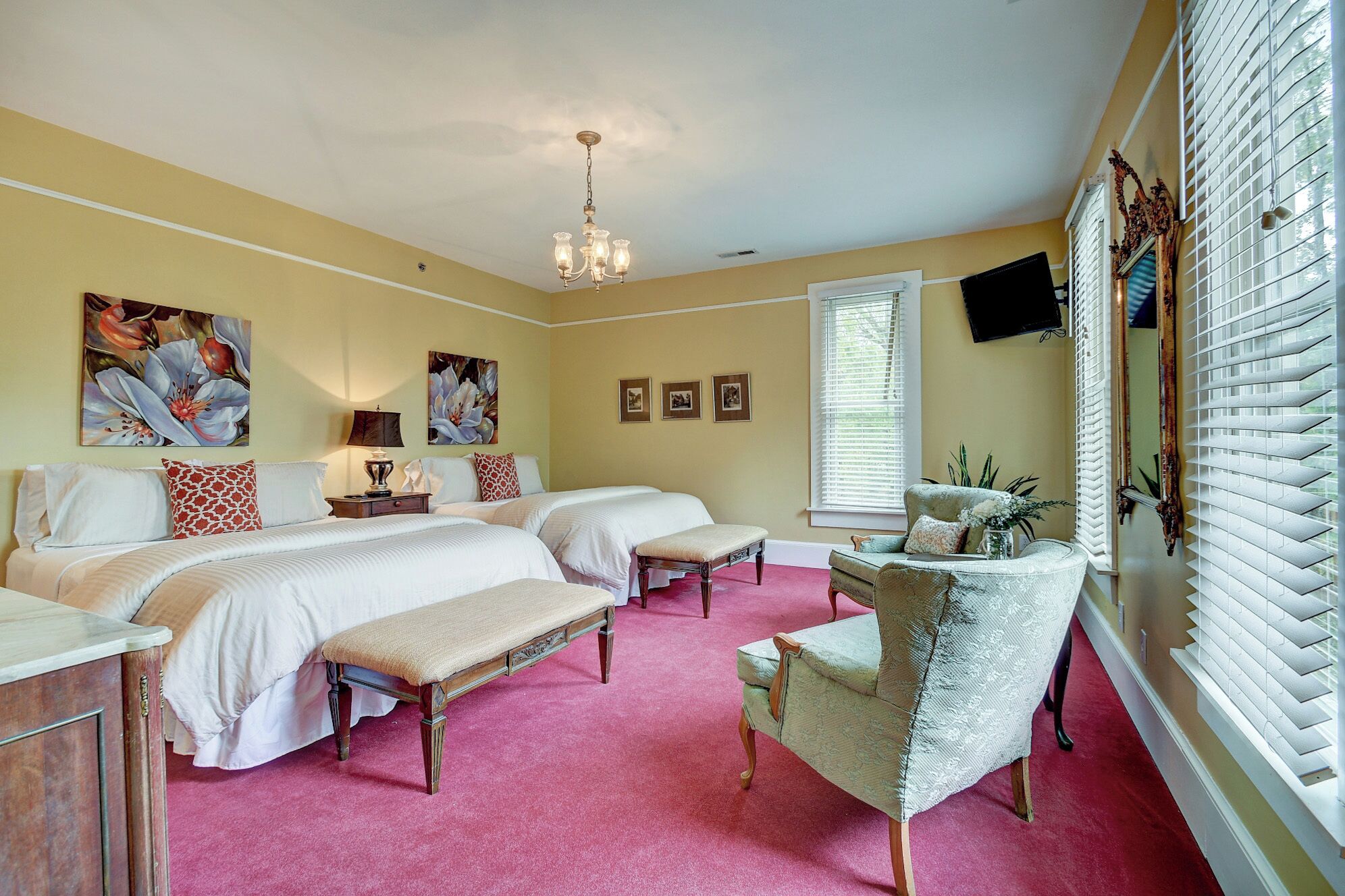 A bedroom with two beds, a bench, a chair, a chandelier, and a large window with blinds.