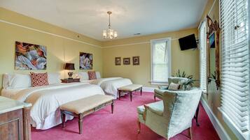 A bedroom with two beds, a bench, a chair, a chandelier, and a large window with blinds.