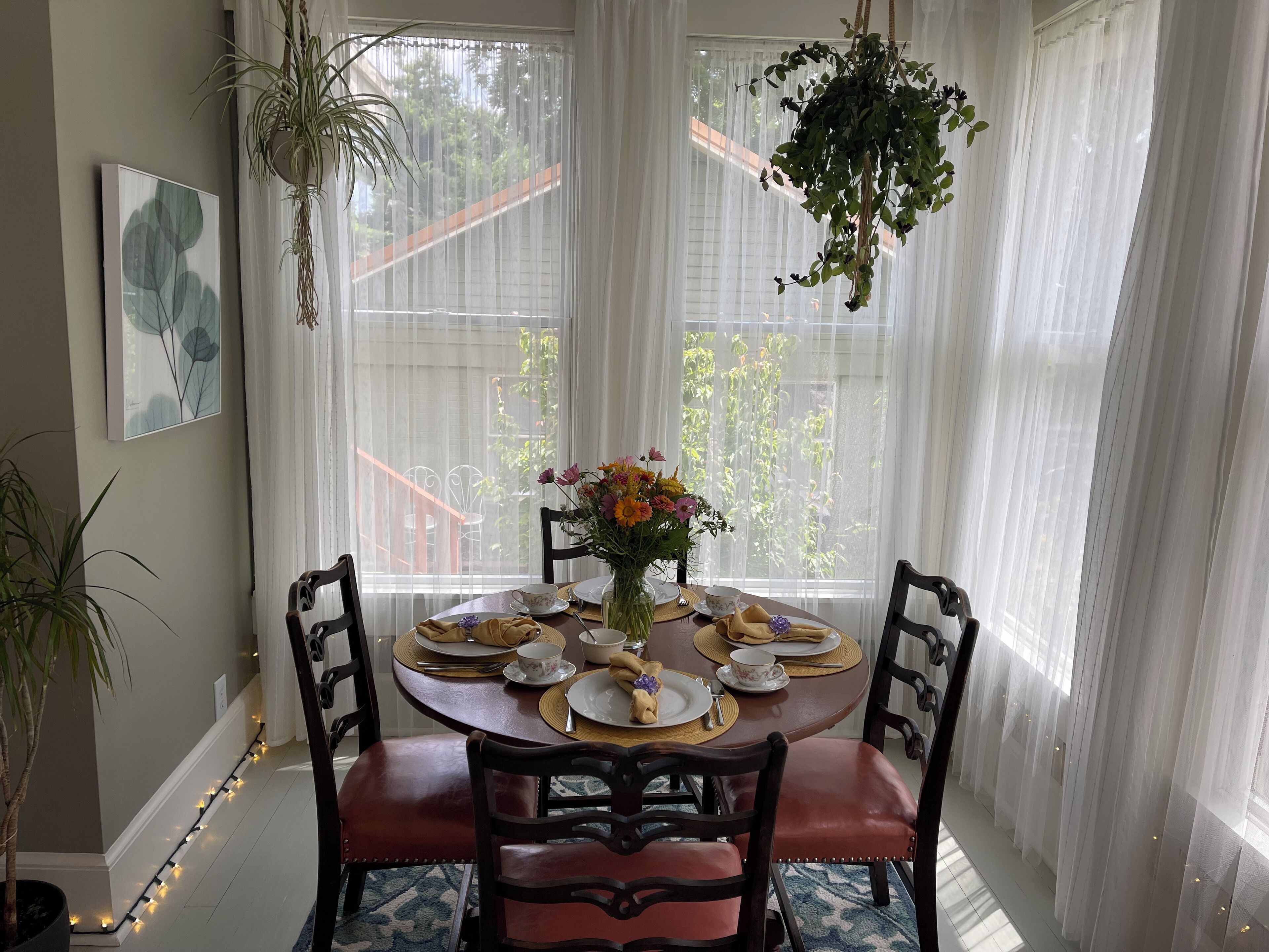 Un comedor con una mesa redonda dispuesta para cuatro personas, rodeada de sillas. Hay una planta colgante sobre la mesa y una ventana con cortinas traslúcidas detrás.