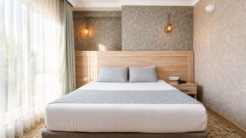 A hotel room with a large bed, two bedside lamps, a nightstand, and a window with sheer curtains.