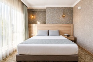 Una habitación de hotel con una cama grande, dos lámparas de noche, una mesita de noche y una ventana con cortinas translúcidas.