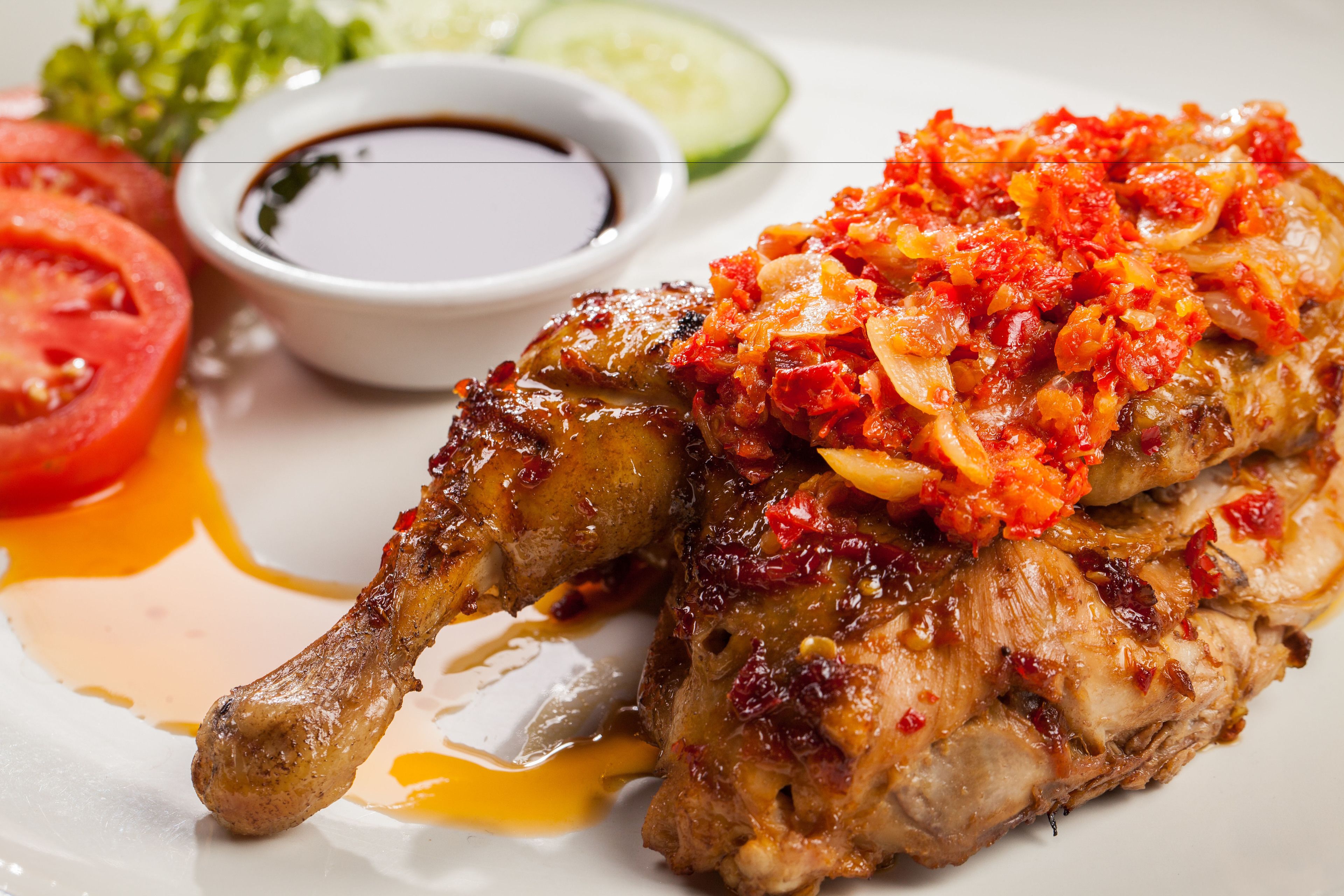 A plate with grilled chicken, a side of sliced tomatoes, a cucumber, and a small bowl of sauce.