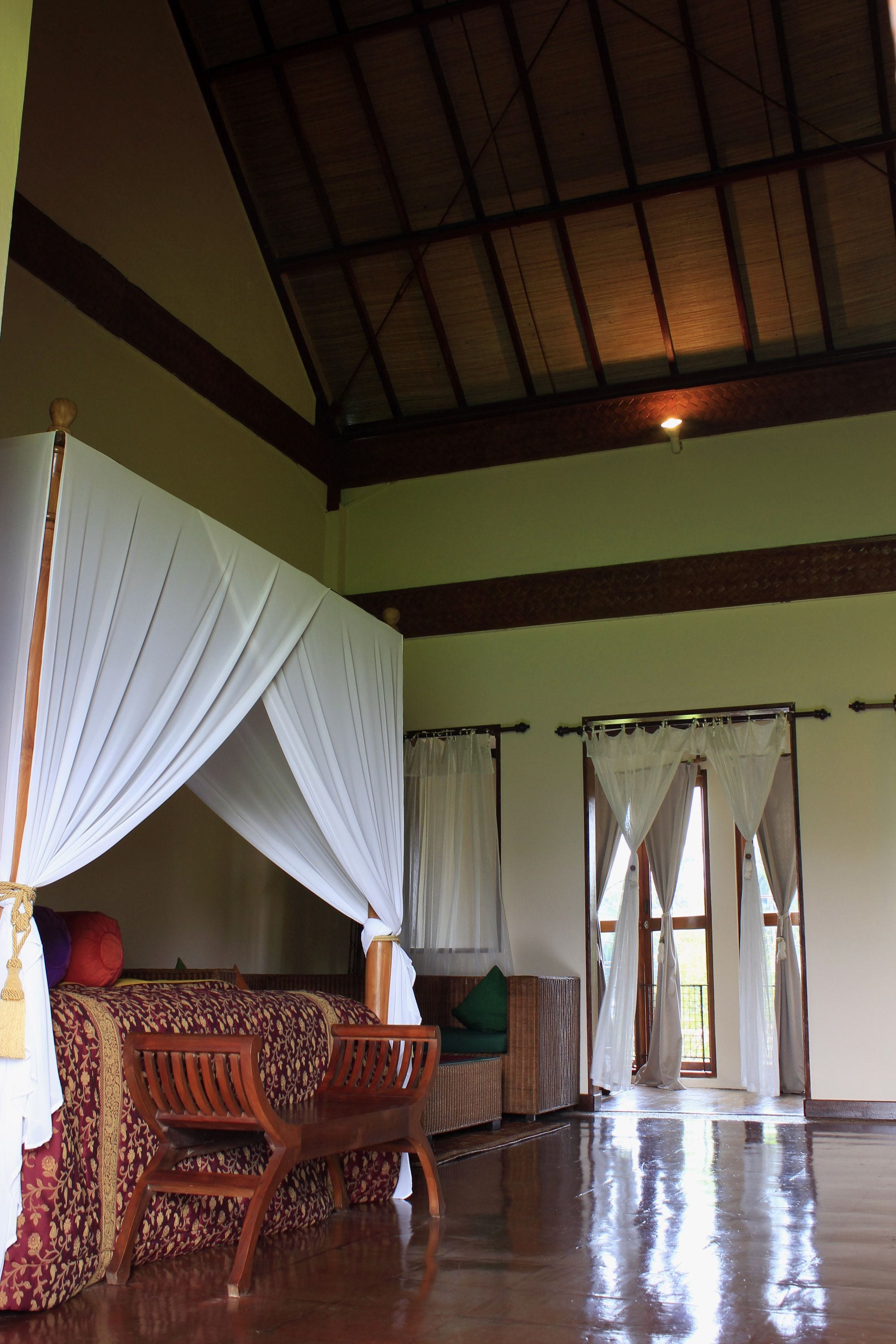 A spacious room with a high ceiling, a bed with a canopy, a wooden bench, and a large window with curtains.