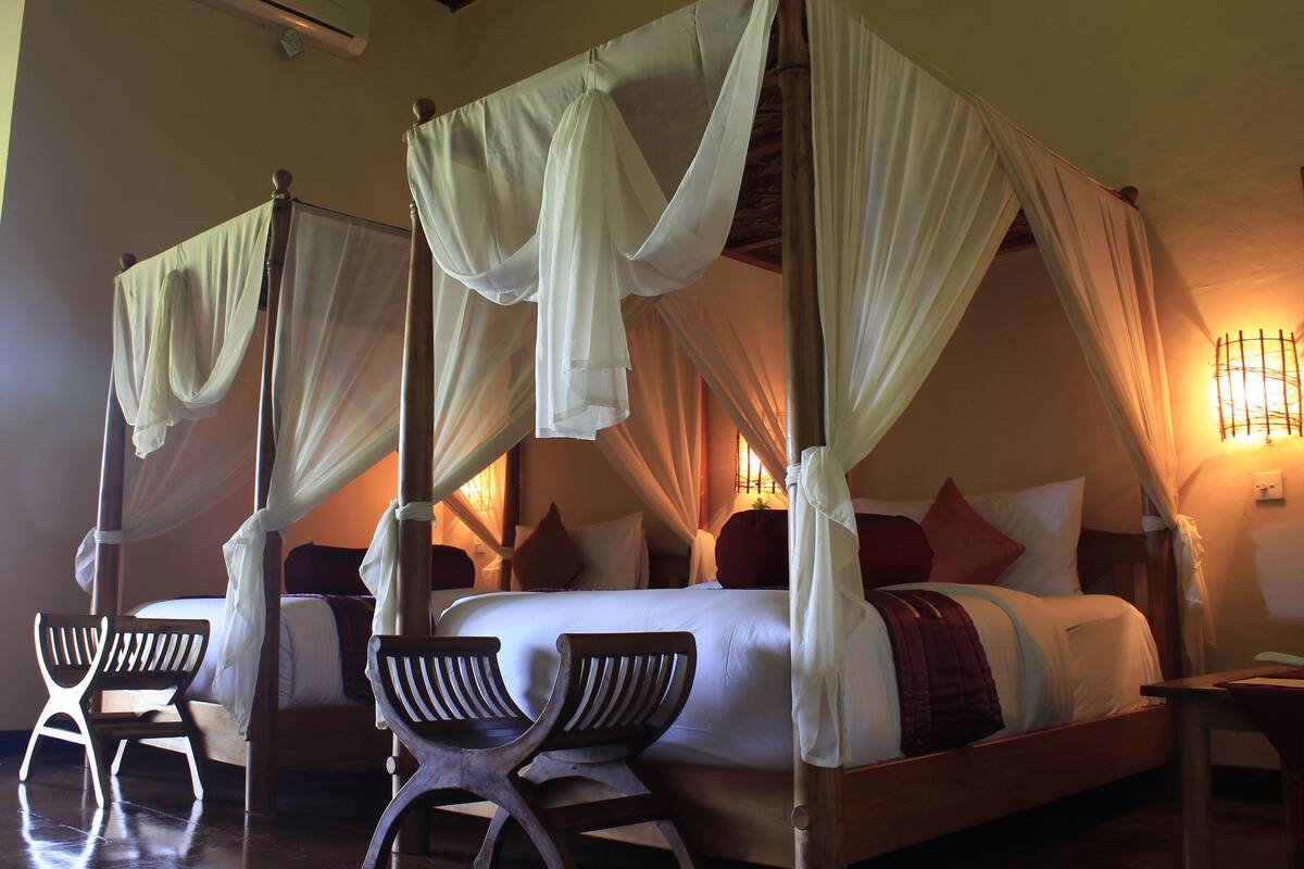 A four-poster canopy bed with white linens and a red throw blanket, two wooden chairs, and a bedside table with a lamp.