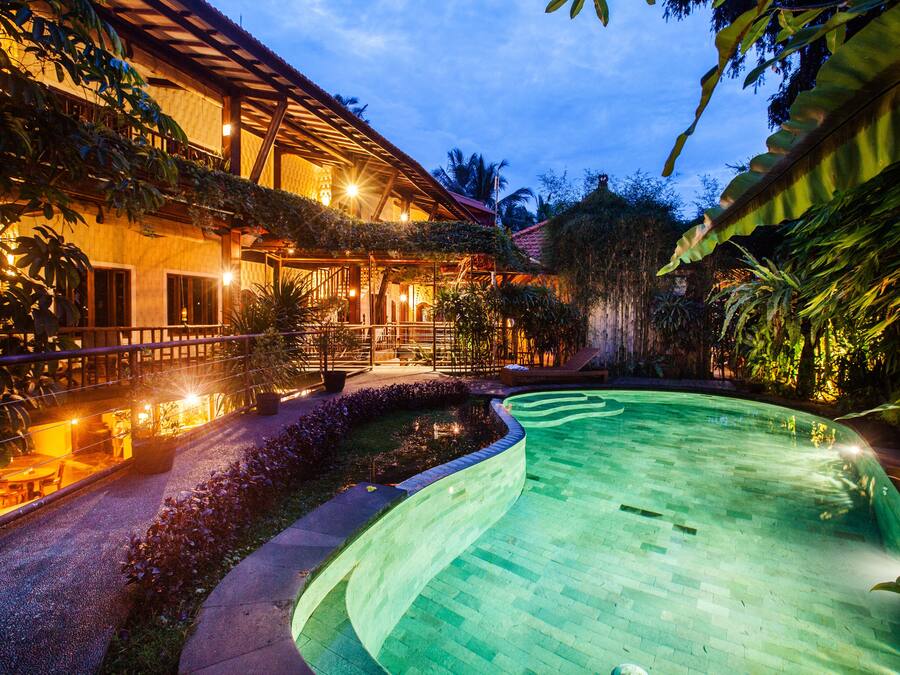 A pool area with a curved edge, surrounded by lush greenery and a building with multiple windows and balconies.