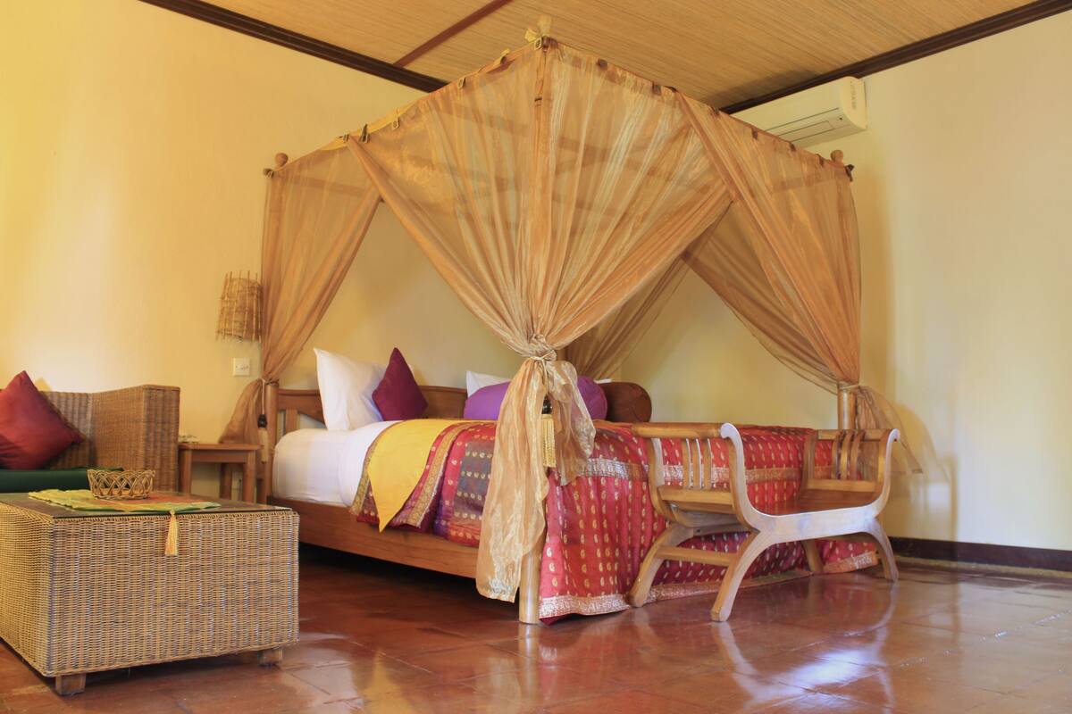 A room with a four-poster bed, a wooden chair, a small table, and a wicker sofa.