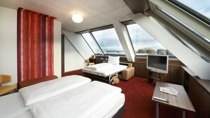 Habitación de hotel moderna con una cama grande, un escritorio, un televisor y vistas de la ciudad a través de amplias ventanas.