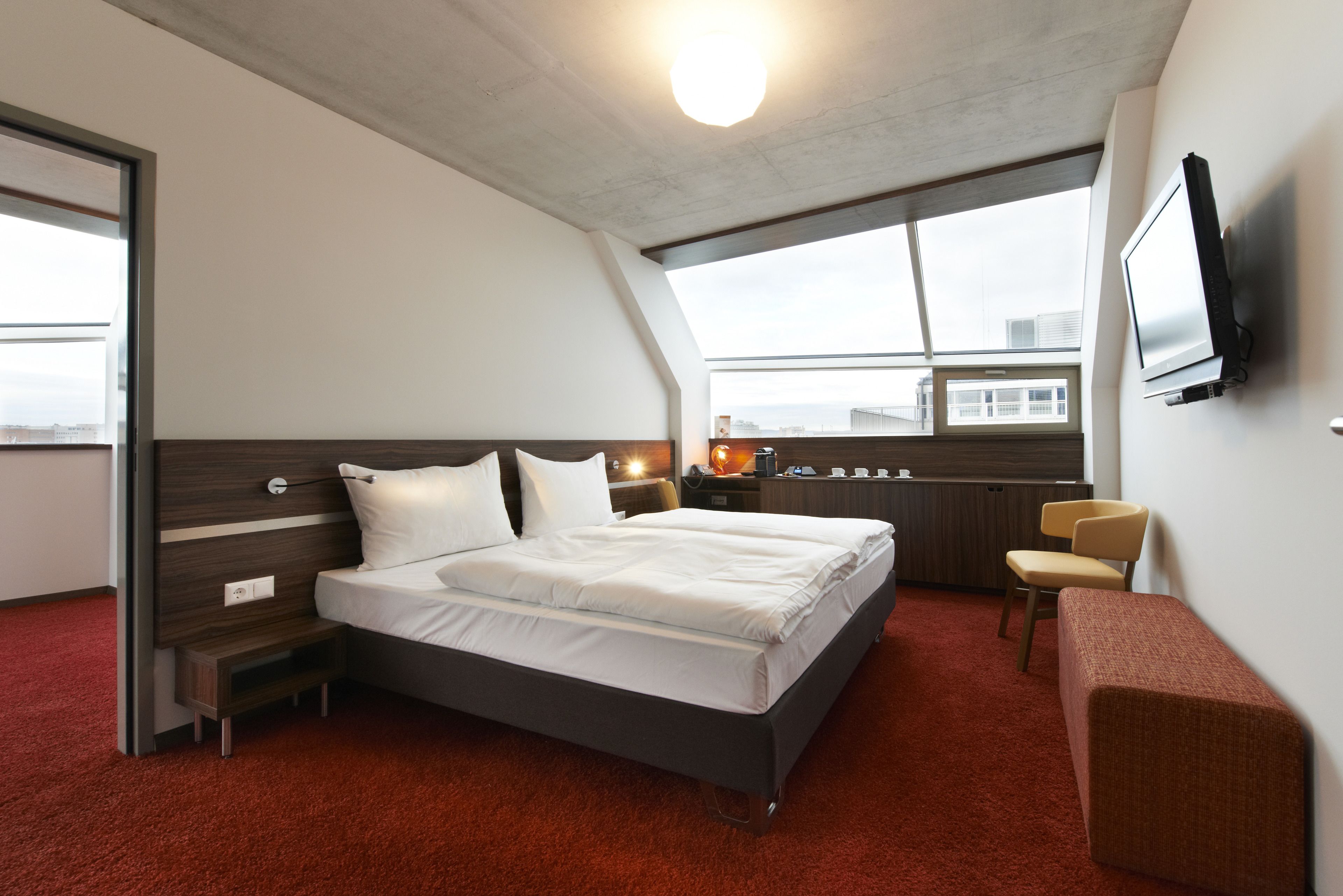 A modern hotel room with a large bed, a desk, a chair, a TV, and a window with a city view.