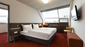 A modern hotel room with a large bed, a desk, a chair, a TV, and a window with a city view.