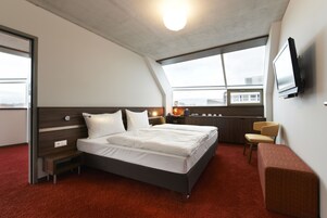 Quarto de hotel moderno com uma cama grande, uma mesa, uma cadeira, uma televisão e uma janela com vista para a cidade.