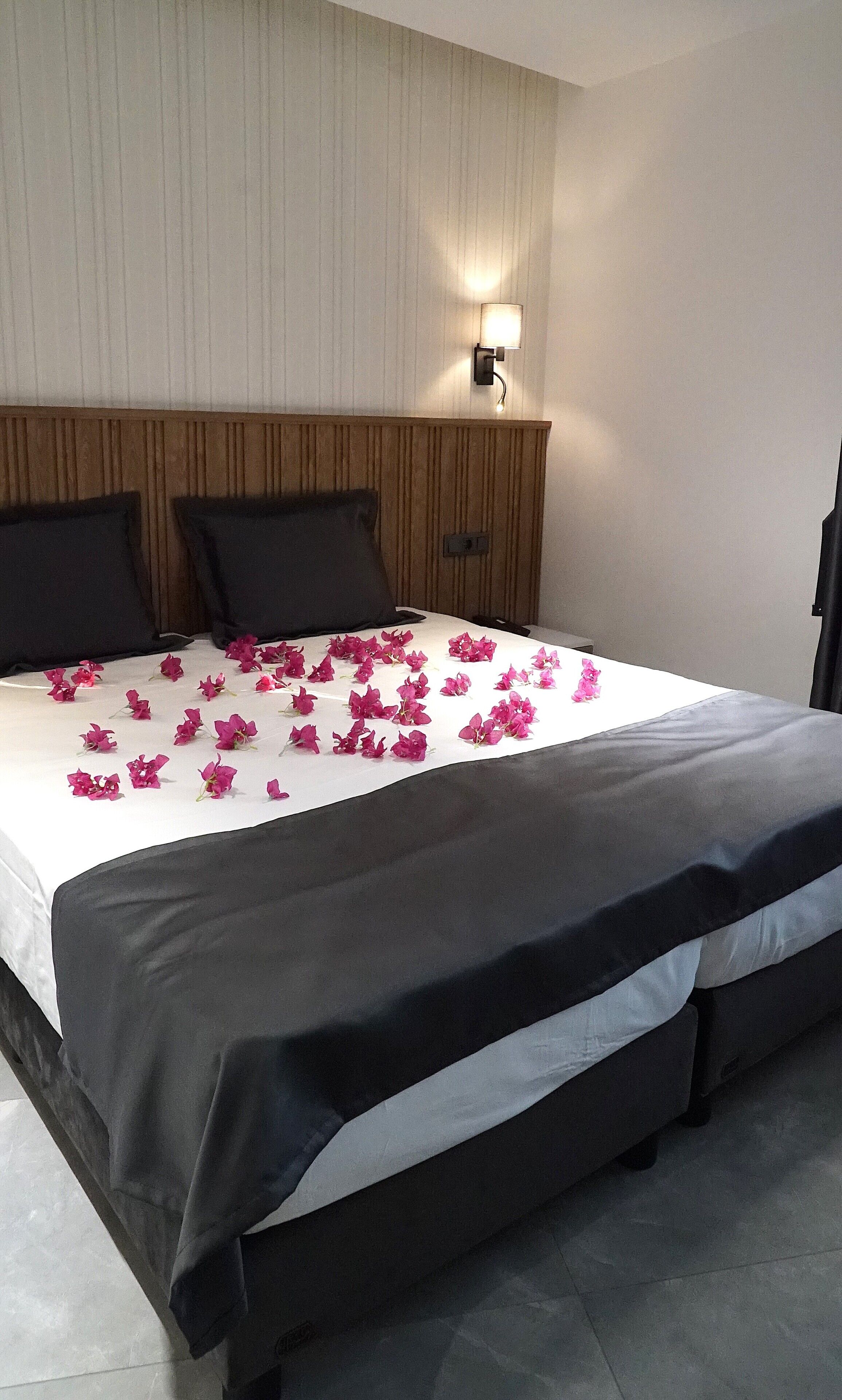 Quarto de hotel com uma cama grande, duas almofadas pretas, um painel de cabeceira de madeira e uma luminária fixada na parede.