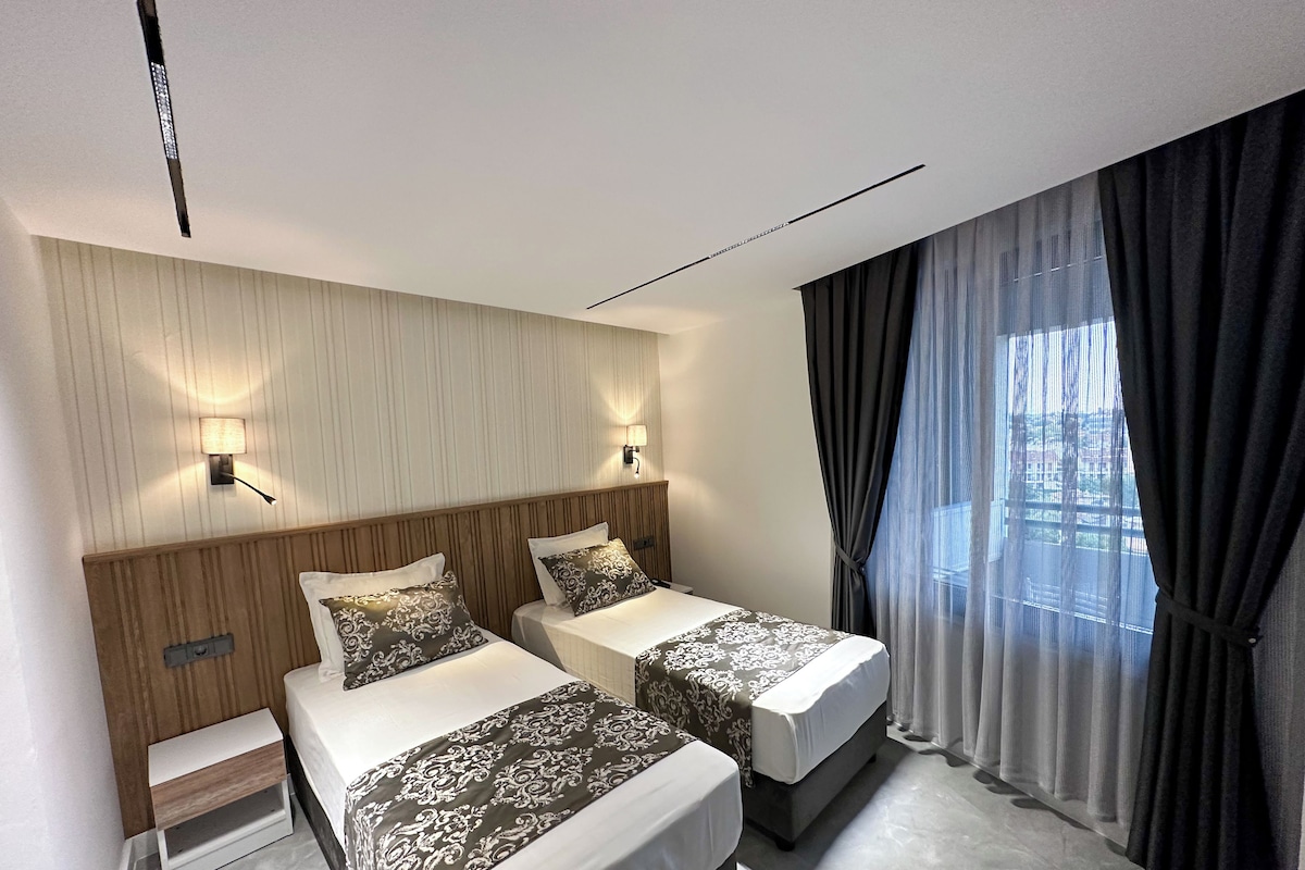 Una habitación de hotel moderna con dos camas, un cabecero de madera, una mesita de noche pequeña y un ventanal grande con cortinas traslúcidas.