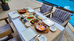 A dining table set with various dishes, including eggs, vegetables, and meats, near a poolside seating area with striped cushions. - Happy Apart (Kusadasi)