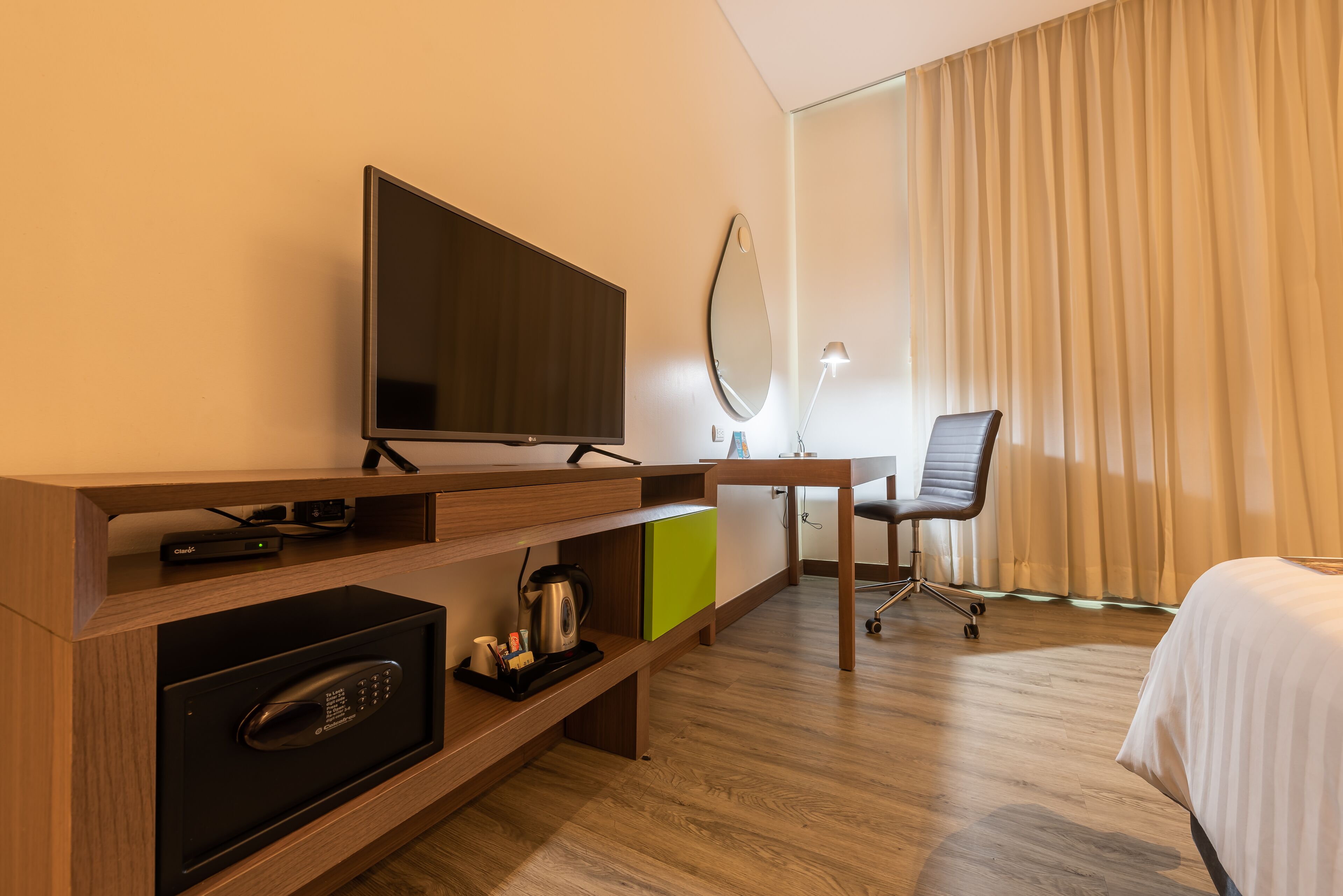 Habitación de hotel con televisión de pantalla plana, escritorio con lámpara, una silla, cama y minibar.