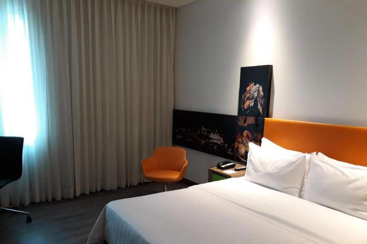 A hotel room with a bed, an orange chair, a nightstand, and a painting on the wall.