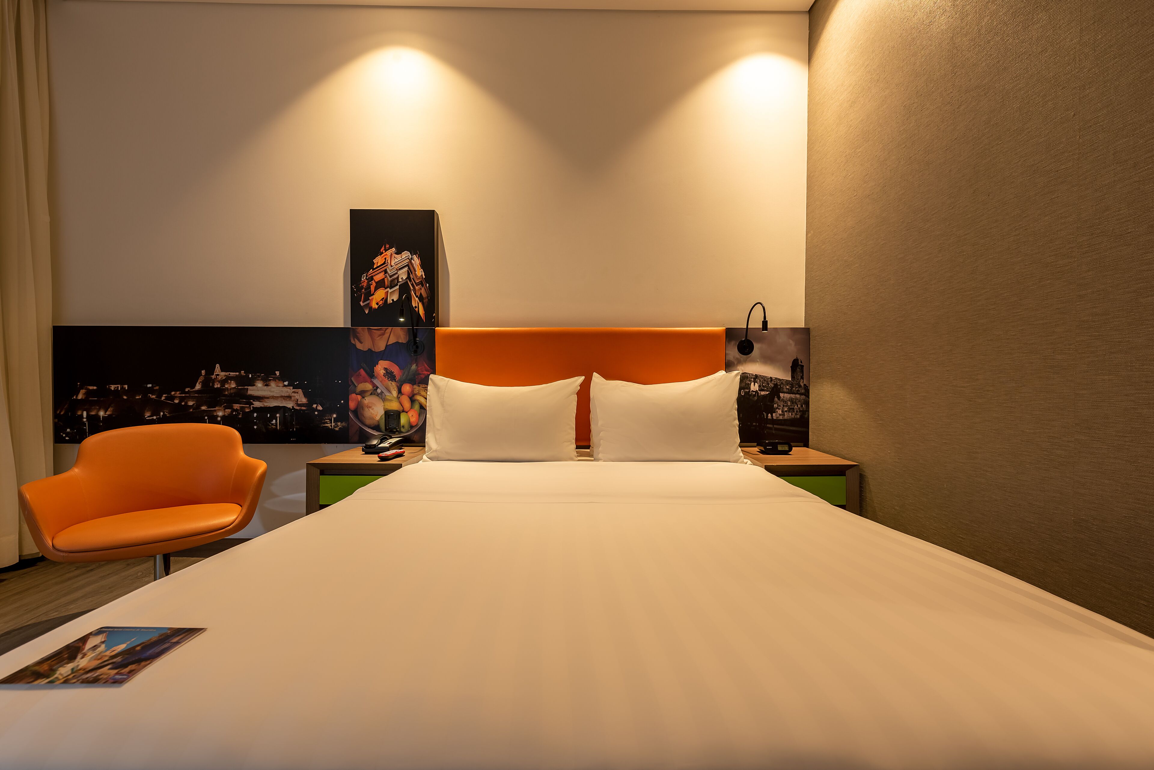 A hotel room with a large bed, a nightstand, a modern orange chair, and a cityscape artwork on the wall.