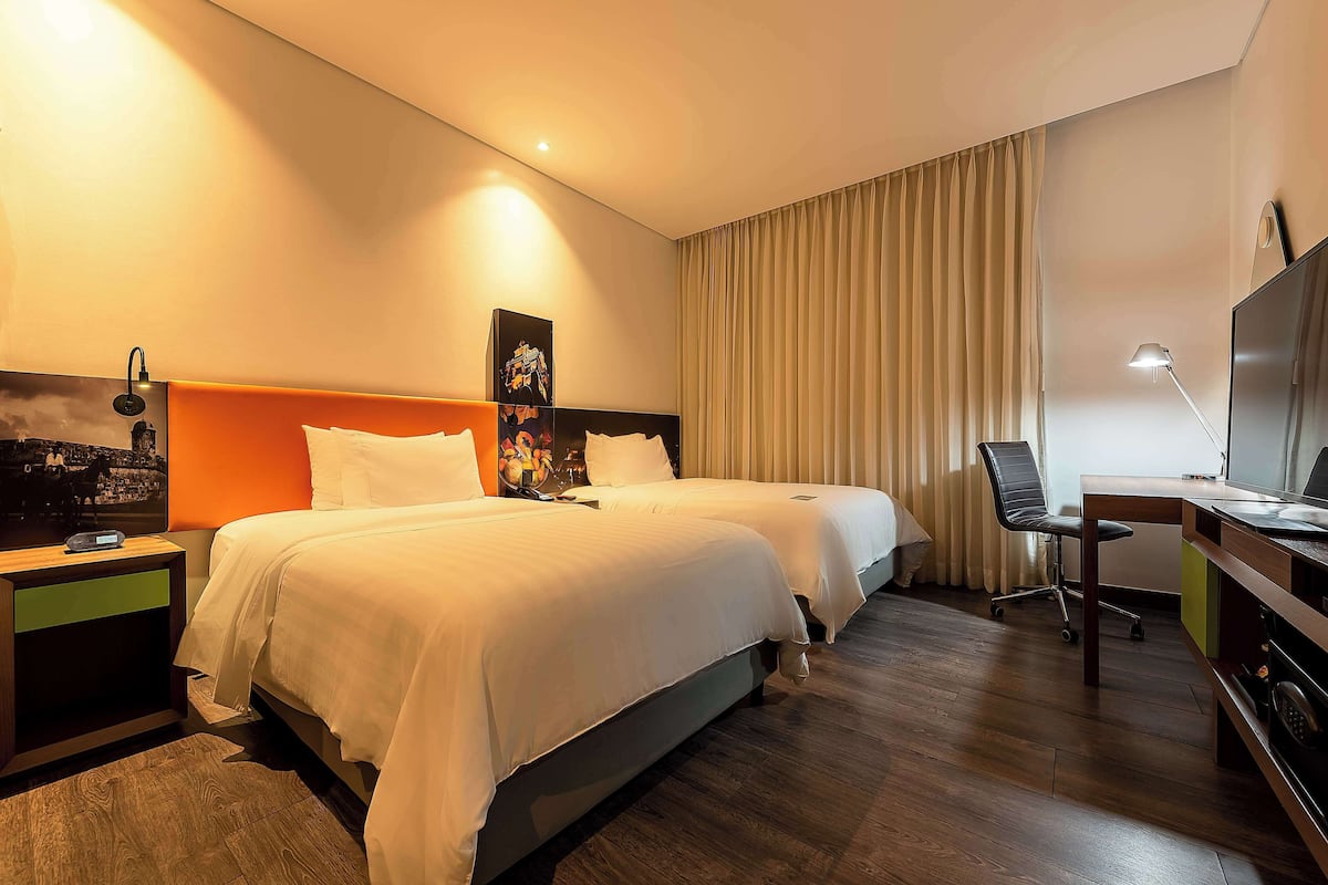 A hotel room with two beds, a desk, a chair, a TV, and a nightstand with a lamp.