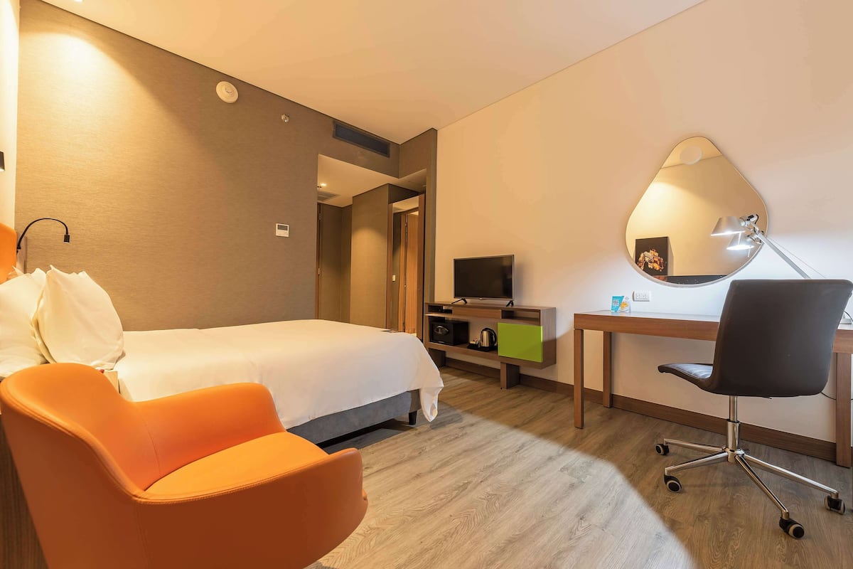 A hotel room with a bed, an orange chair, a desk with a chair, a TV, and a mirror on the wall.