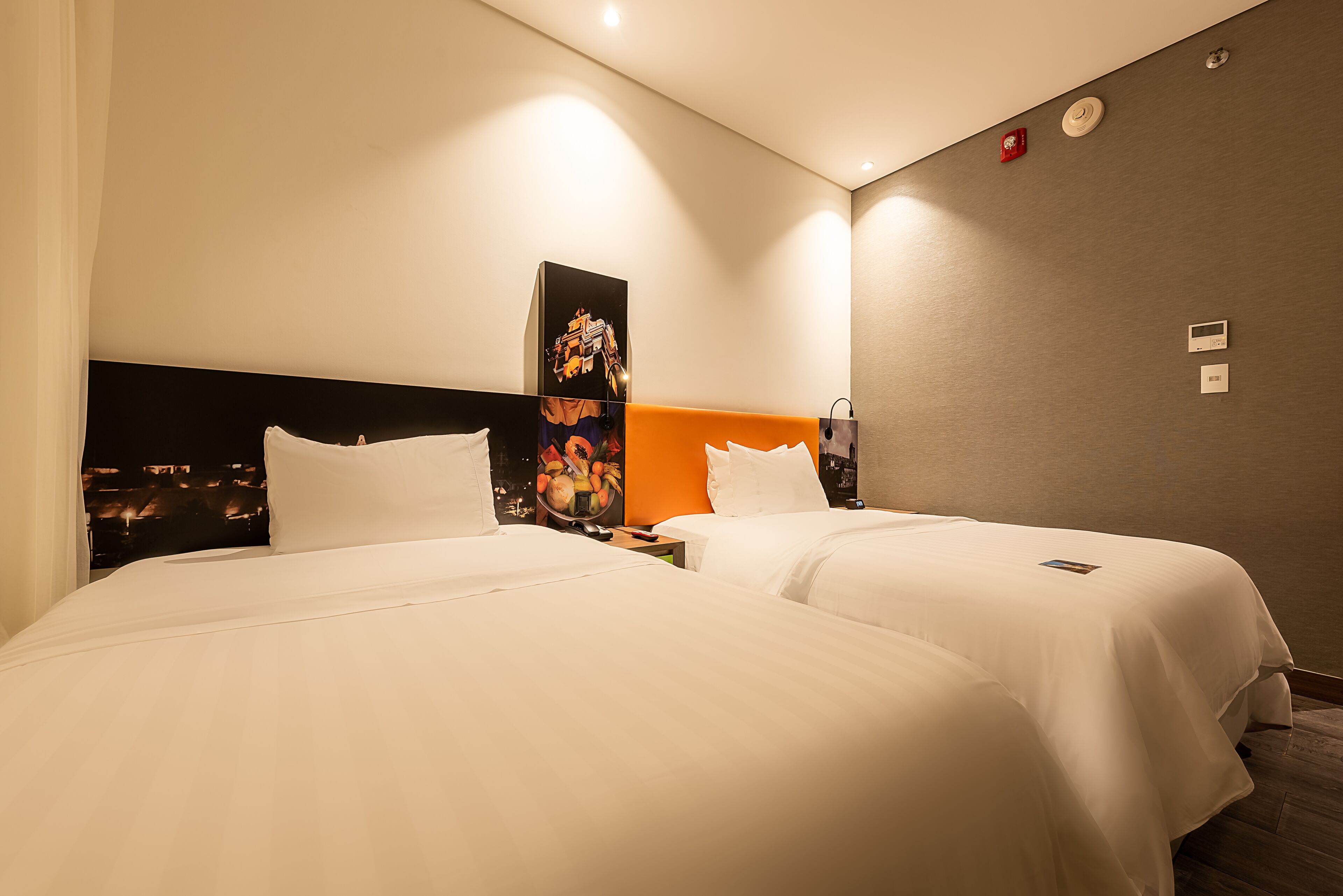A hotel room with two beds, a headboard with artwork, a nightstand, and a wall-mounted control panel.