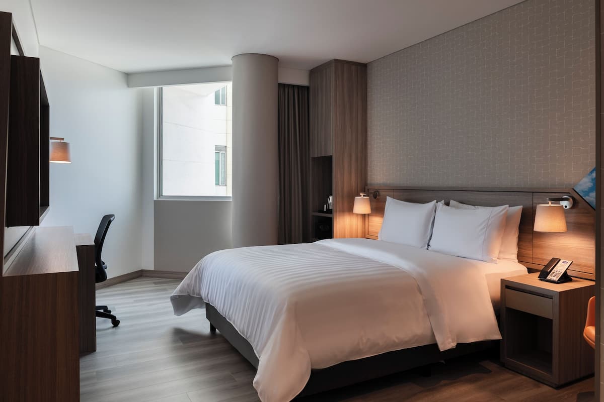 A hotel room with a large bed, a desk, a chair, a telephone, and a window.