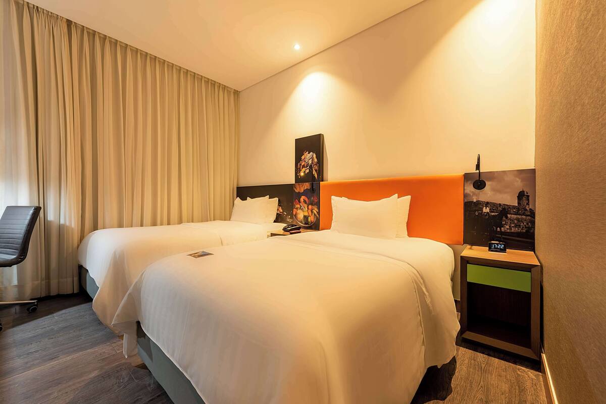 A hotel room with two beds, a chair, a nightstand with a clock, and a wall-mounted picture.