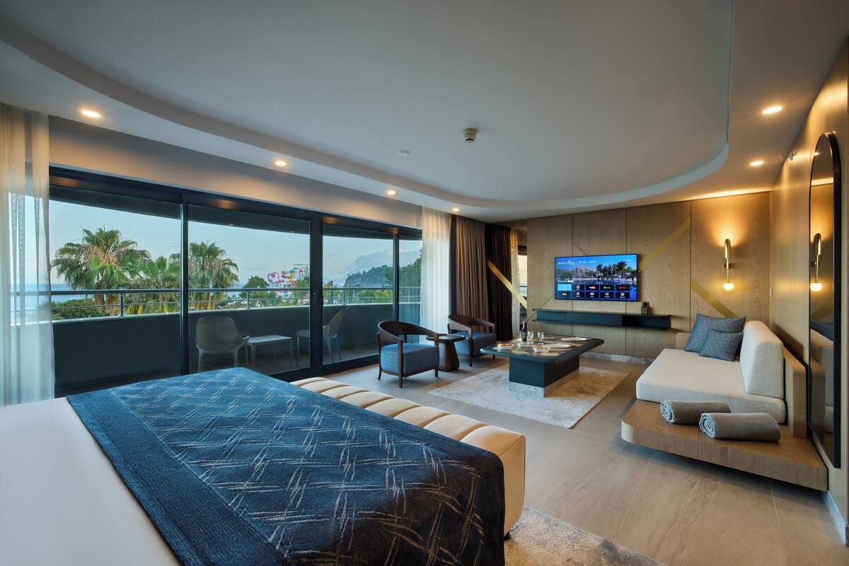 A modern hotel room with a large bed, a sofa, a TV, and a balcony with a view of palm trees and mountains.